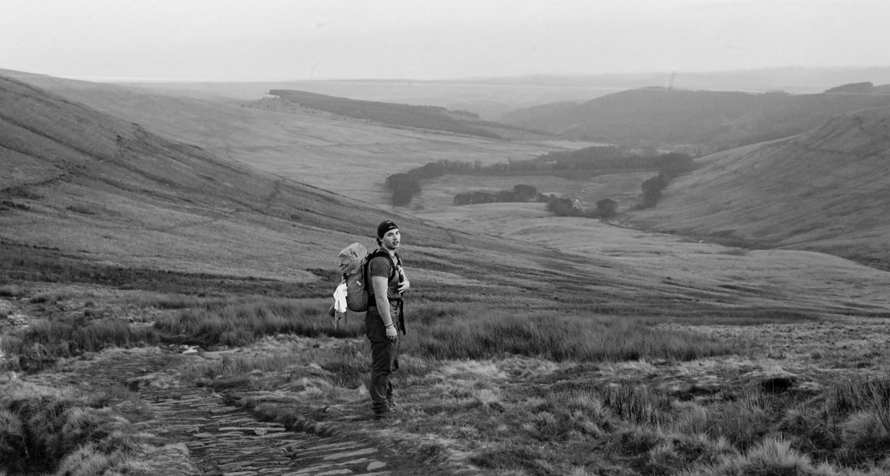 Hiker with a large backpack standing on a path looking across open Brecon Beacons moorland in black and white film