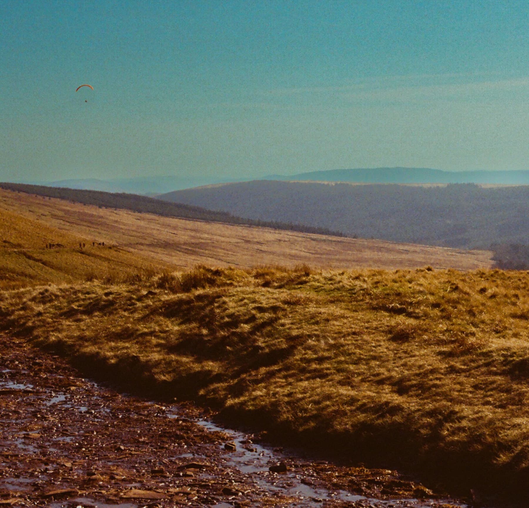 Open moorland with a stream and a distant paraglider in the sky above rolling hills, shot on film