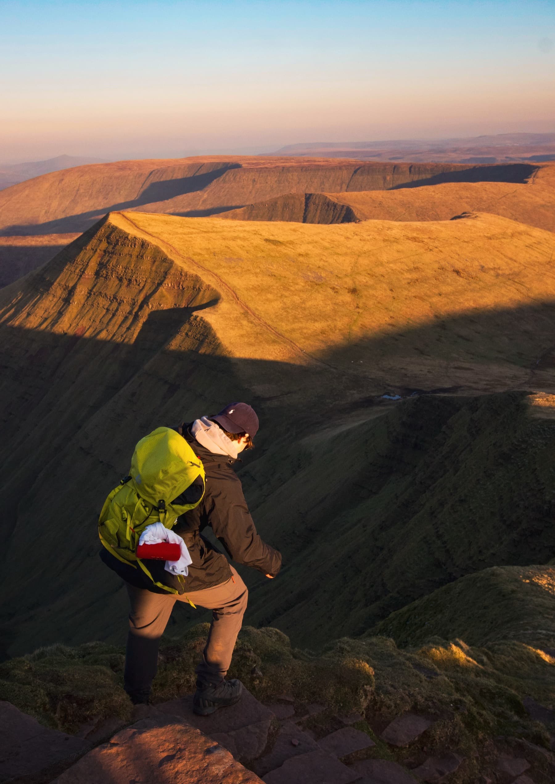 Hiker with green backpack climbing toward the Pen y Fan summit at golden hour with sweeping ridgelines lit by warm light