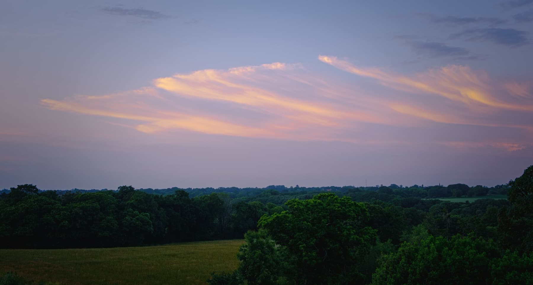 Pink and orange wispy clouds streaking across a pastel sunset sky above a green canopy of trees and fields at twilight