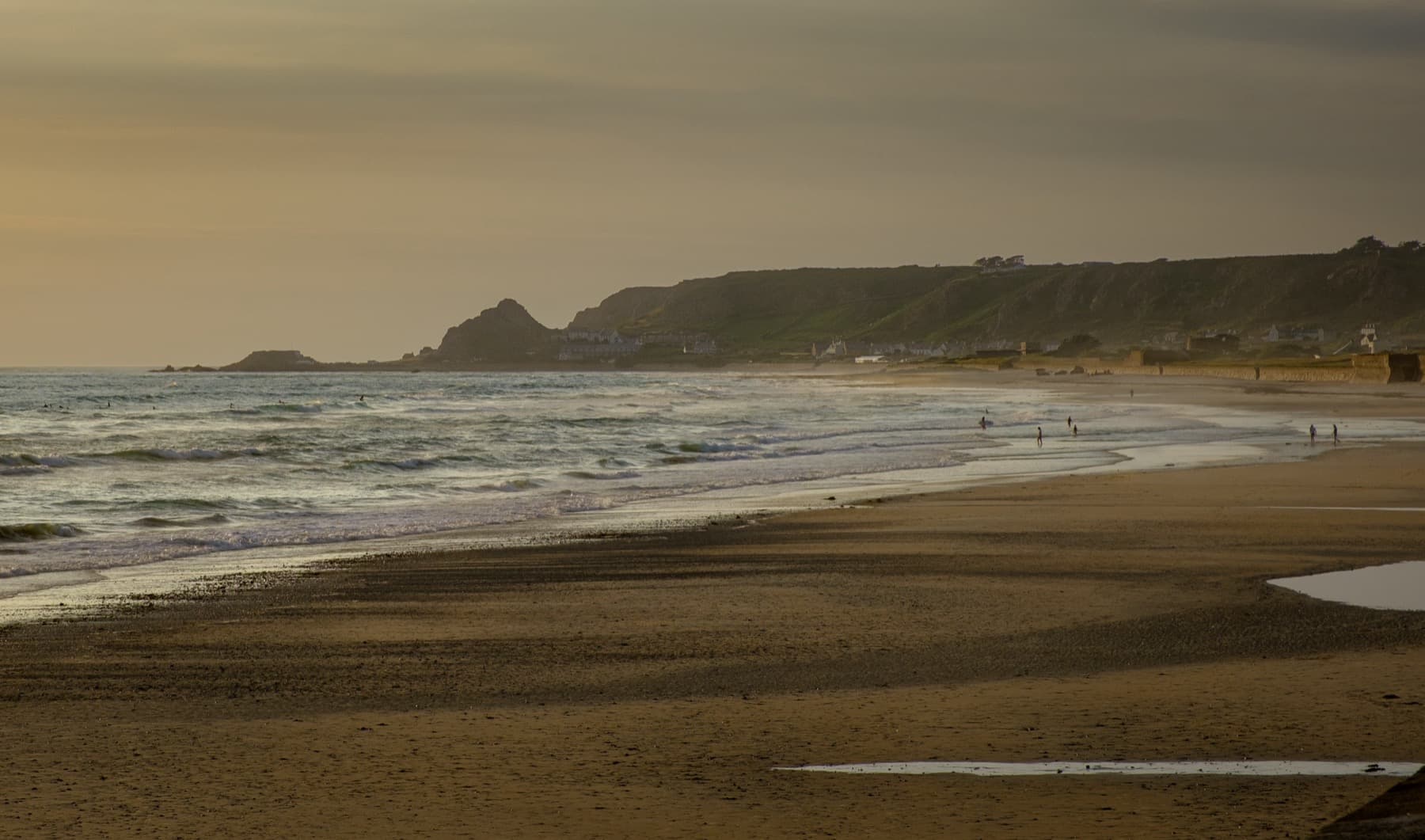 Wide sandy beach at golden hour with distant figures walking along the shoreline, waves breaking and green cliffs rising in the background
