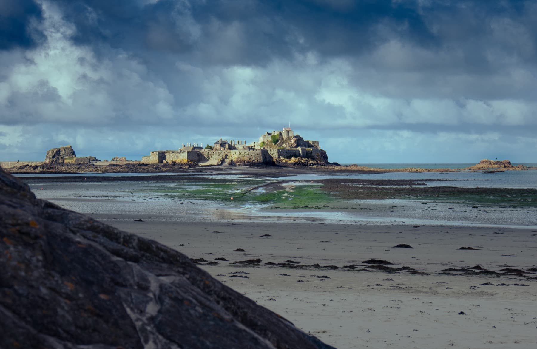 Elizabeth Castle on its rocky tidal island seen across an exposed sandy beach at low tide under dramatic stormy clouds