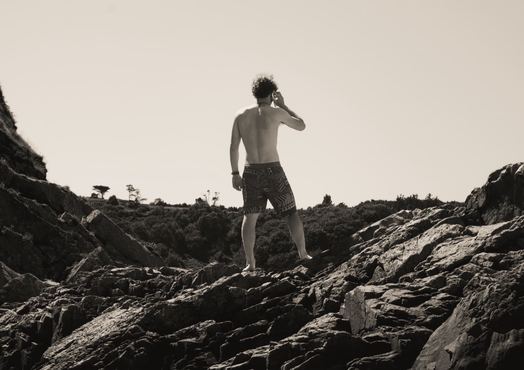 Black and white photograph of a shirtless young man standing on jagged coastal rocks, seen from behind, looking out toward scrubland
