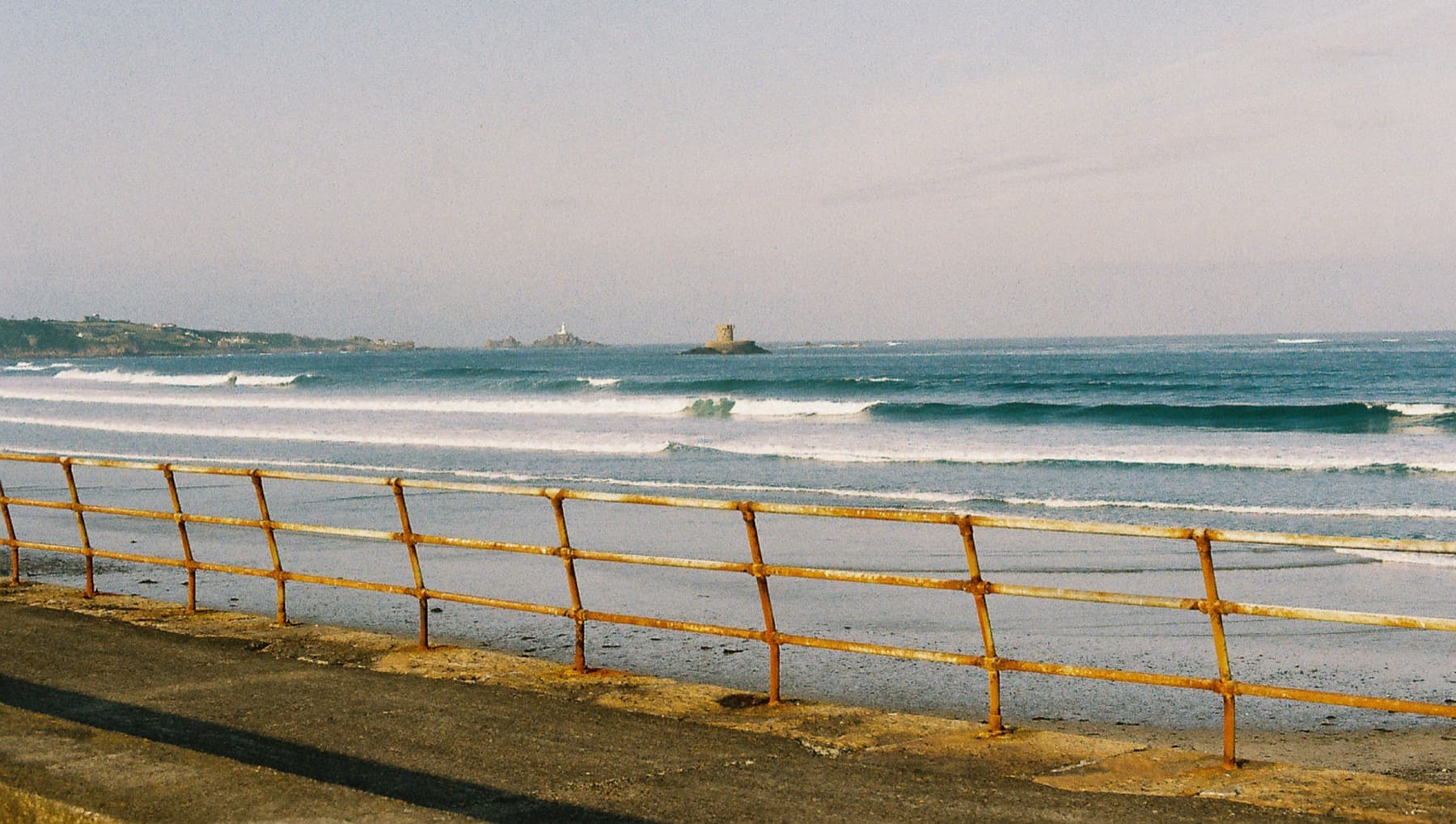 Rows of turquoise waves seen through a weathered orange metal railing along a seaside promenade, with a distant tower on the horizon