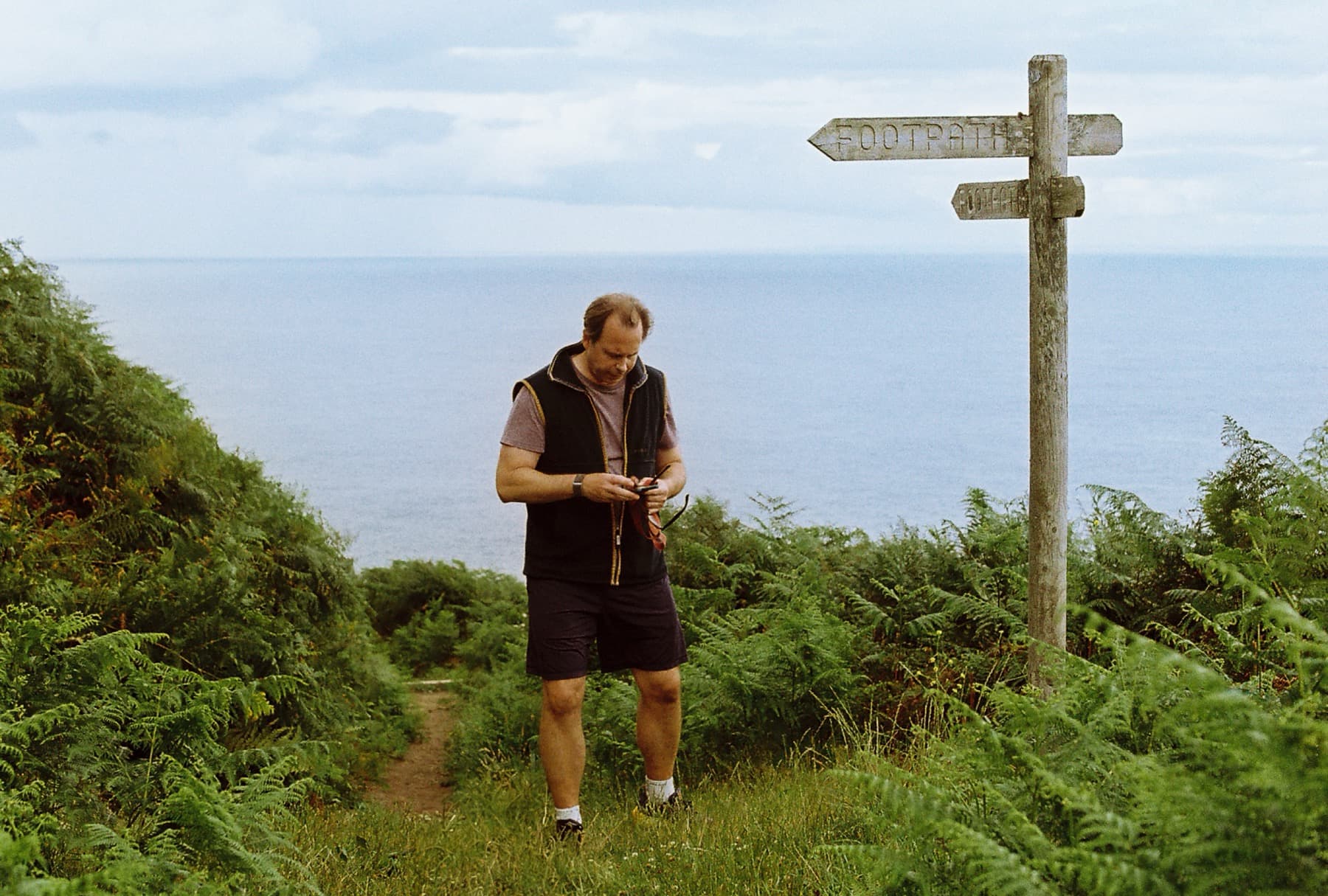 Man checking his phone beside a wooden footpath signpost on a fern-lined coastal cliff path with a calm blue sea in the background