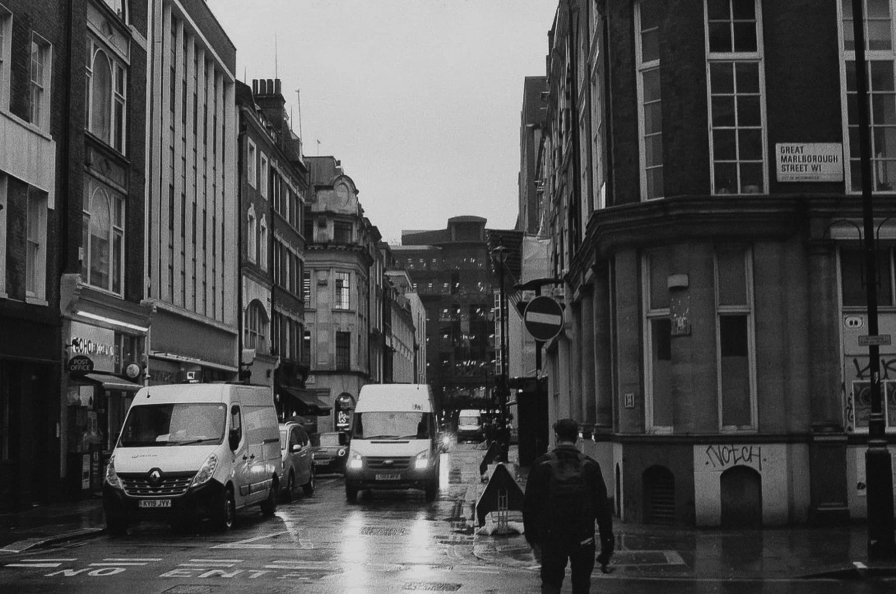 Pedestrians walking on rain-soaked Great Marlborough Street in London with parked vans and historic buildings in black and white