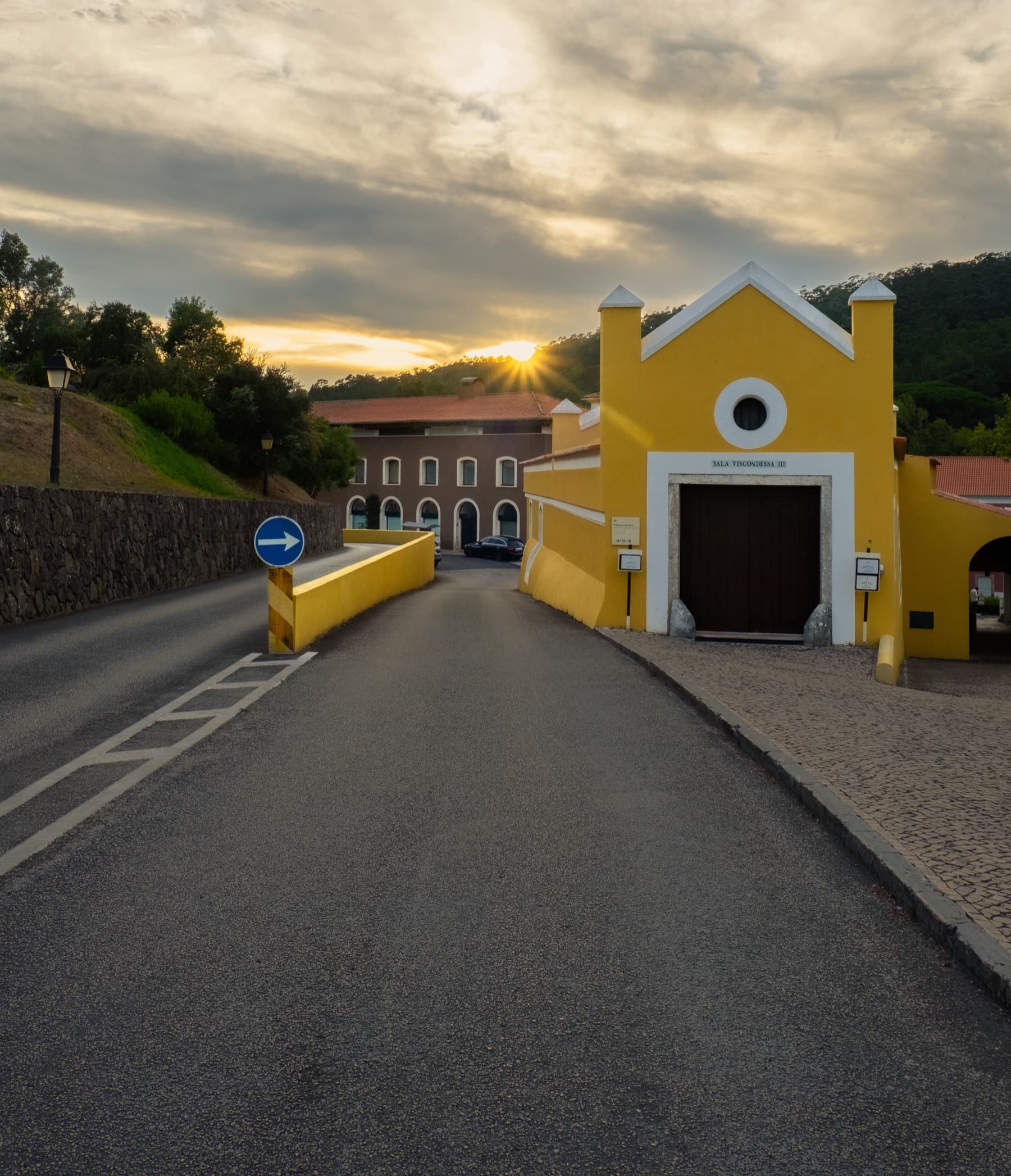 Yellow Portuguese building with arched doorway at the end of a road as the sun sets behind green hills