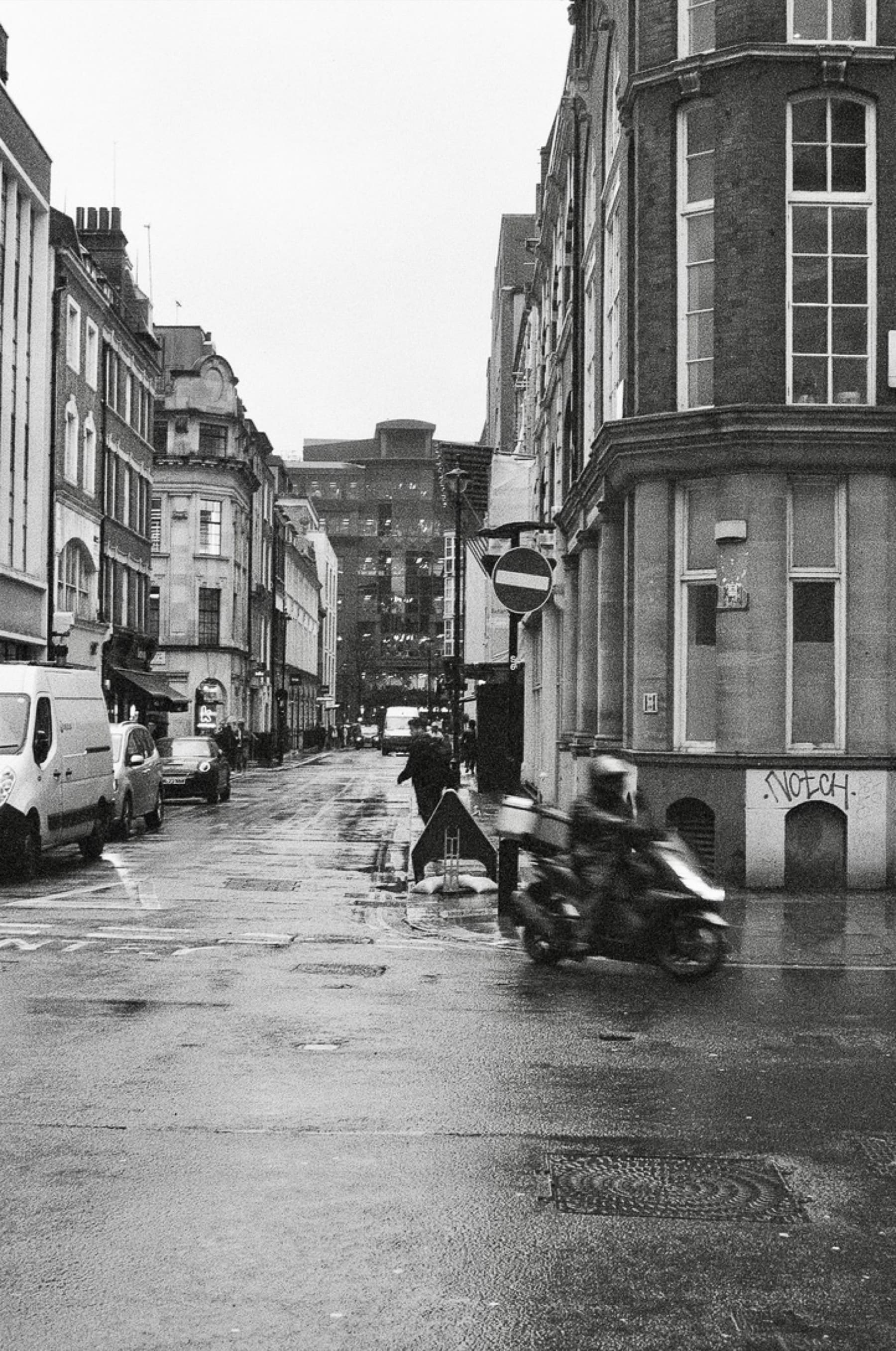 Motion-blurred motorcyclist rounding a corner on a rainy London street in black and white with graffiti on the building