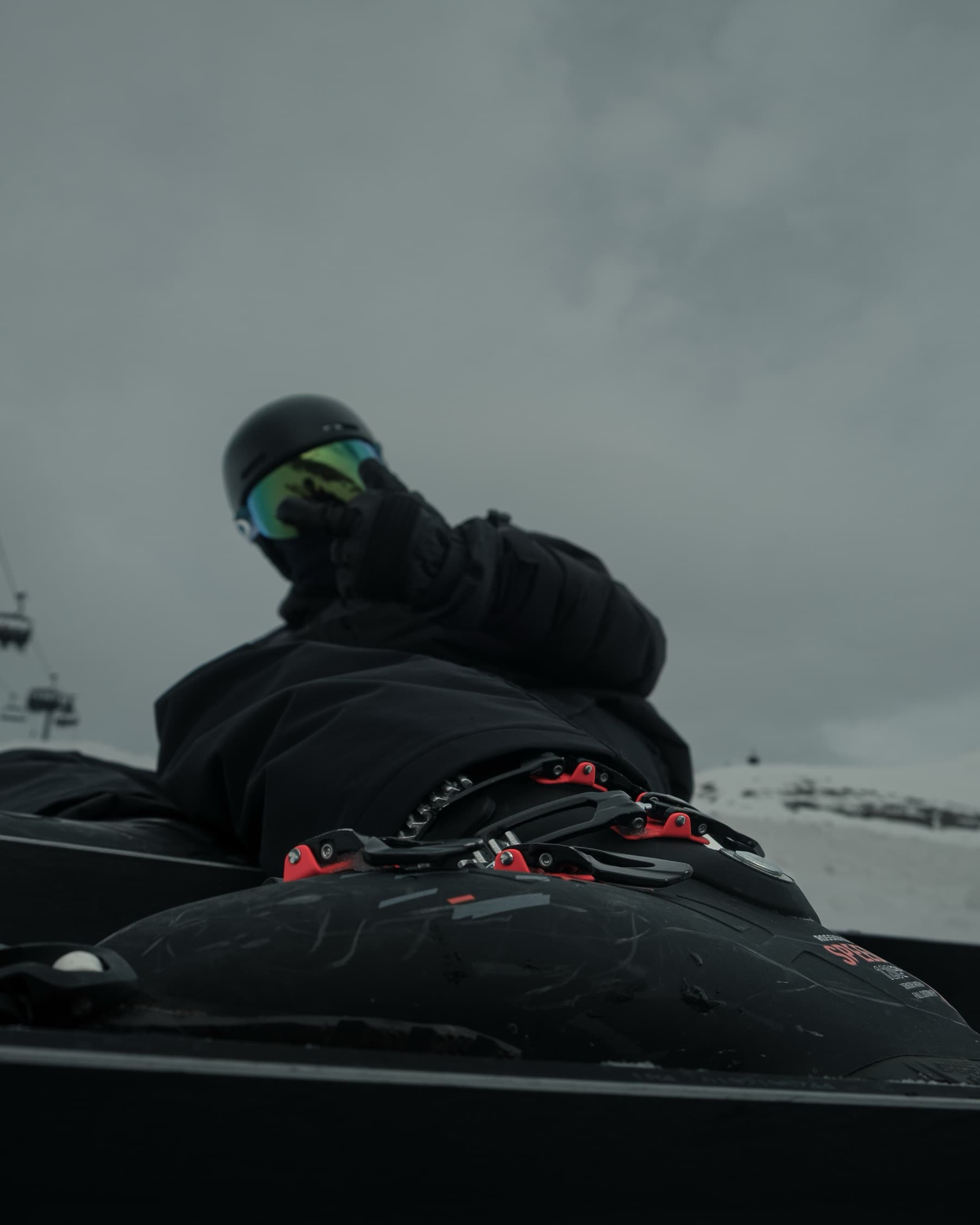 Low-angle view of a skier resting on the snow, shot from ski boot level showing black ski boots with red buckles and reflective goggles against an overcast sky