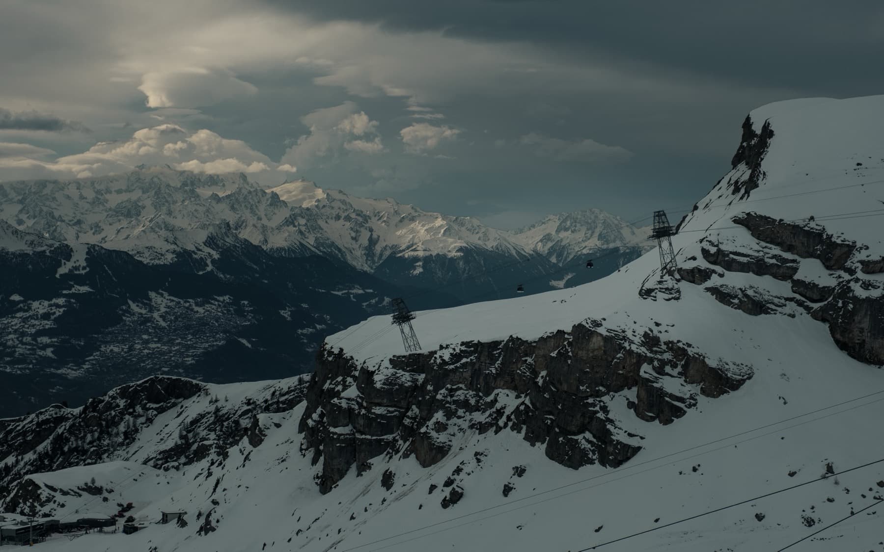 Cable car pylons and gondola on a snow-covered cliff edge with exposed rocky outcrops, overlooking a vast alpine valley under stormy skies
