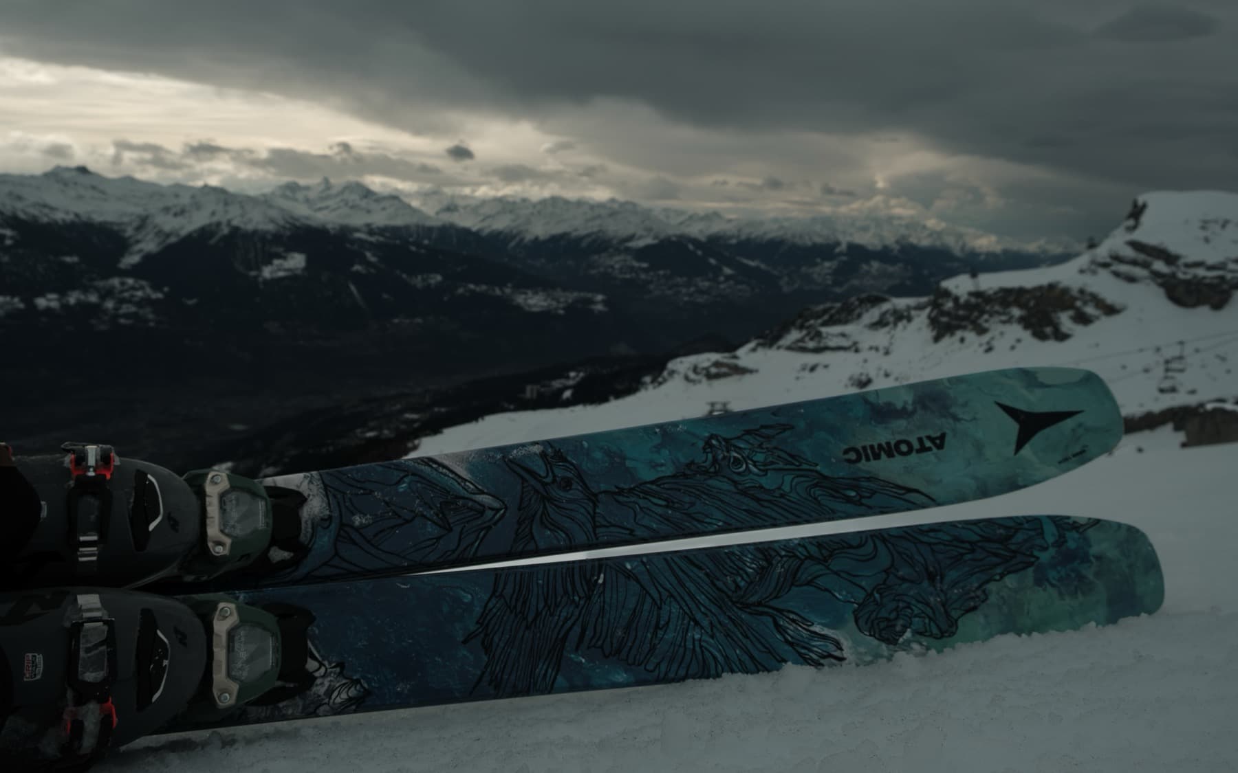 Close-up of Atomic skis with artistic blue and black graphics resting in snow, with a moody mountain panorama and overcast sky behind