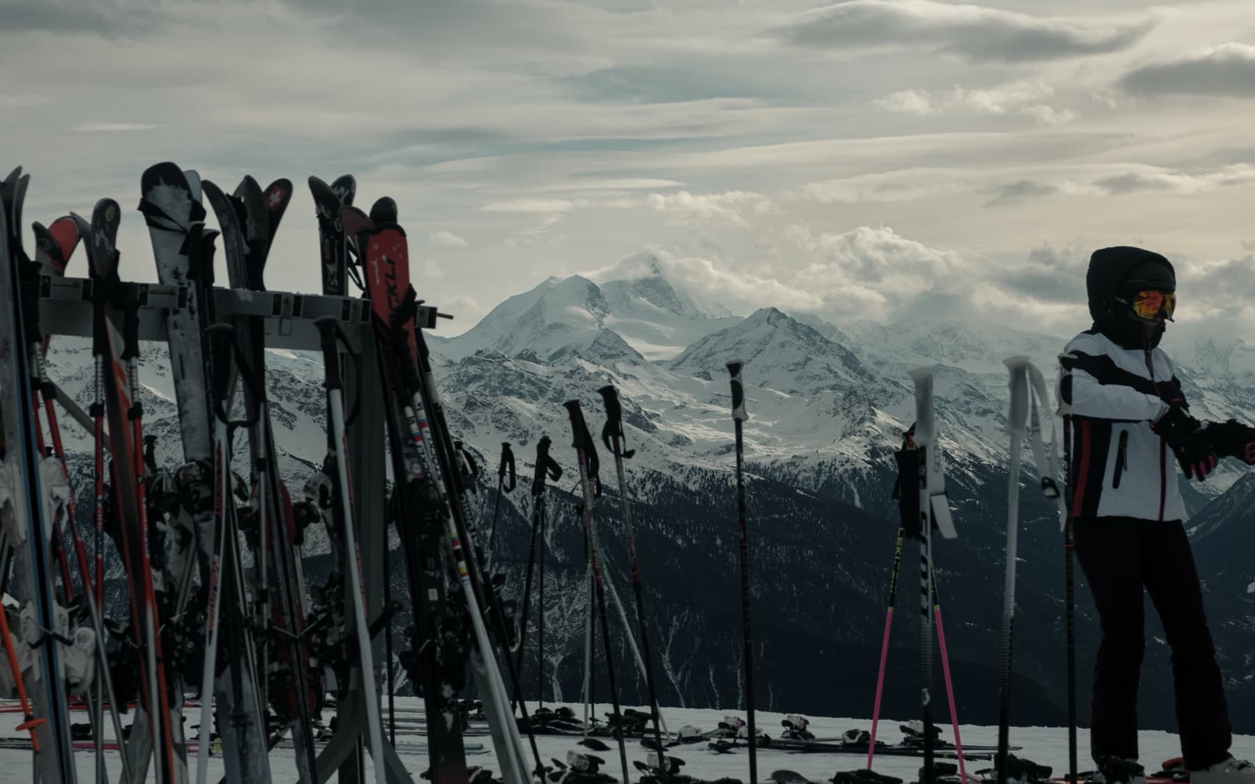 Rows of skis and poles stored in a rack at a mountain restaurant, with a skier standing beside them looking toward distant snow-covered peaks under cloudy skies