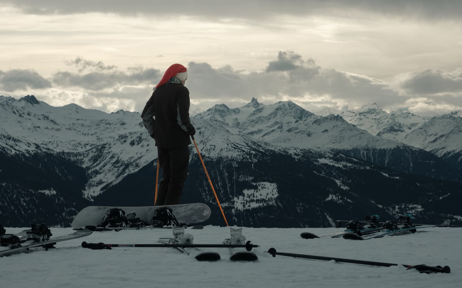 Snowboarder wearing a Santa hat standing on a summit looking out over a vast range of snow-covered peaks under moody clouds, boards and skis laid in the snow