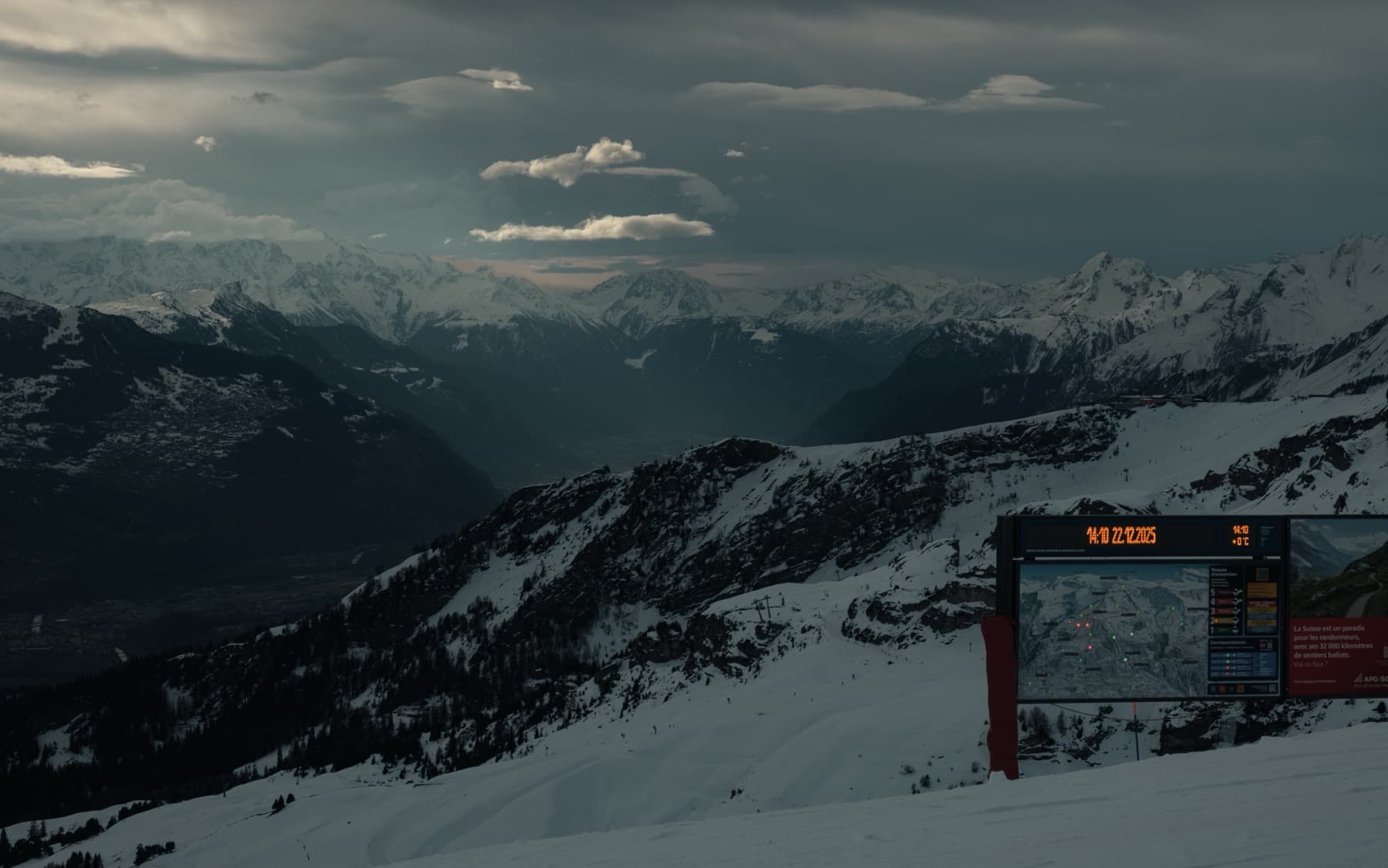 Ski resort information board with digital clock and piste map at the top of a run, panoramic view of snow-covered mountains and deep valley behind