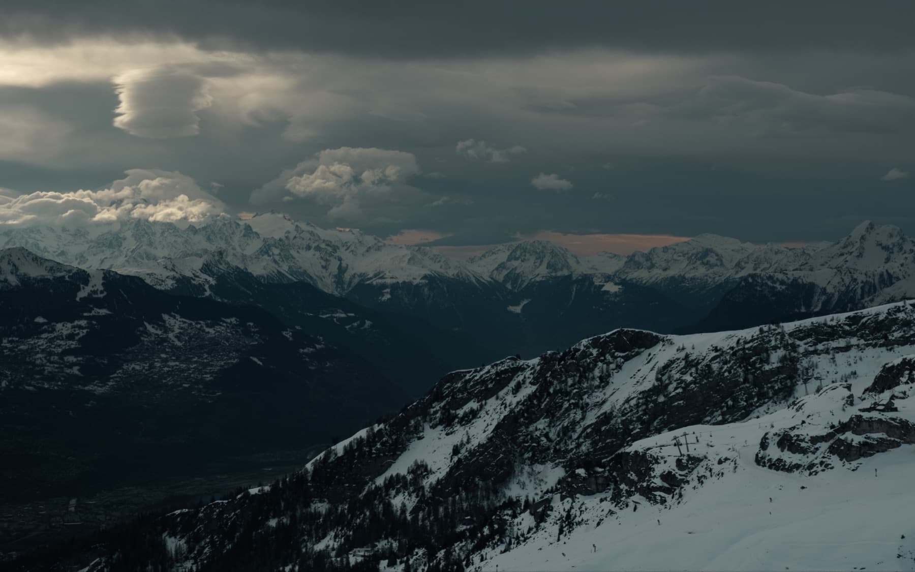 Sweeping mountain valley view under dramatic dark storm clouds with patches of warm light breaking through onto distant snow-covered peaks
