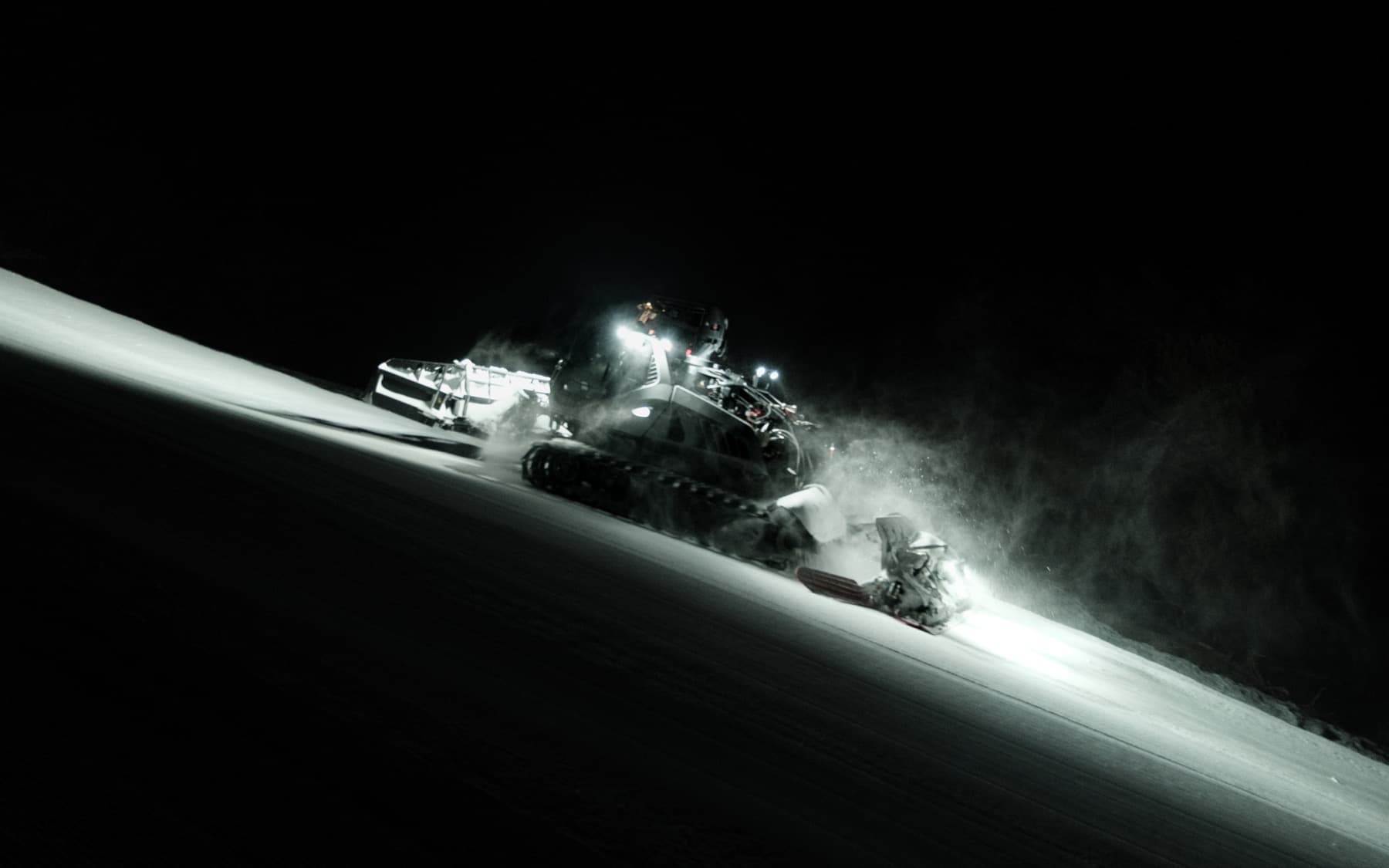 Piste basher grooming machine working at night on a steep ski slope, headlights illuminating the snow and powder spray
