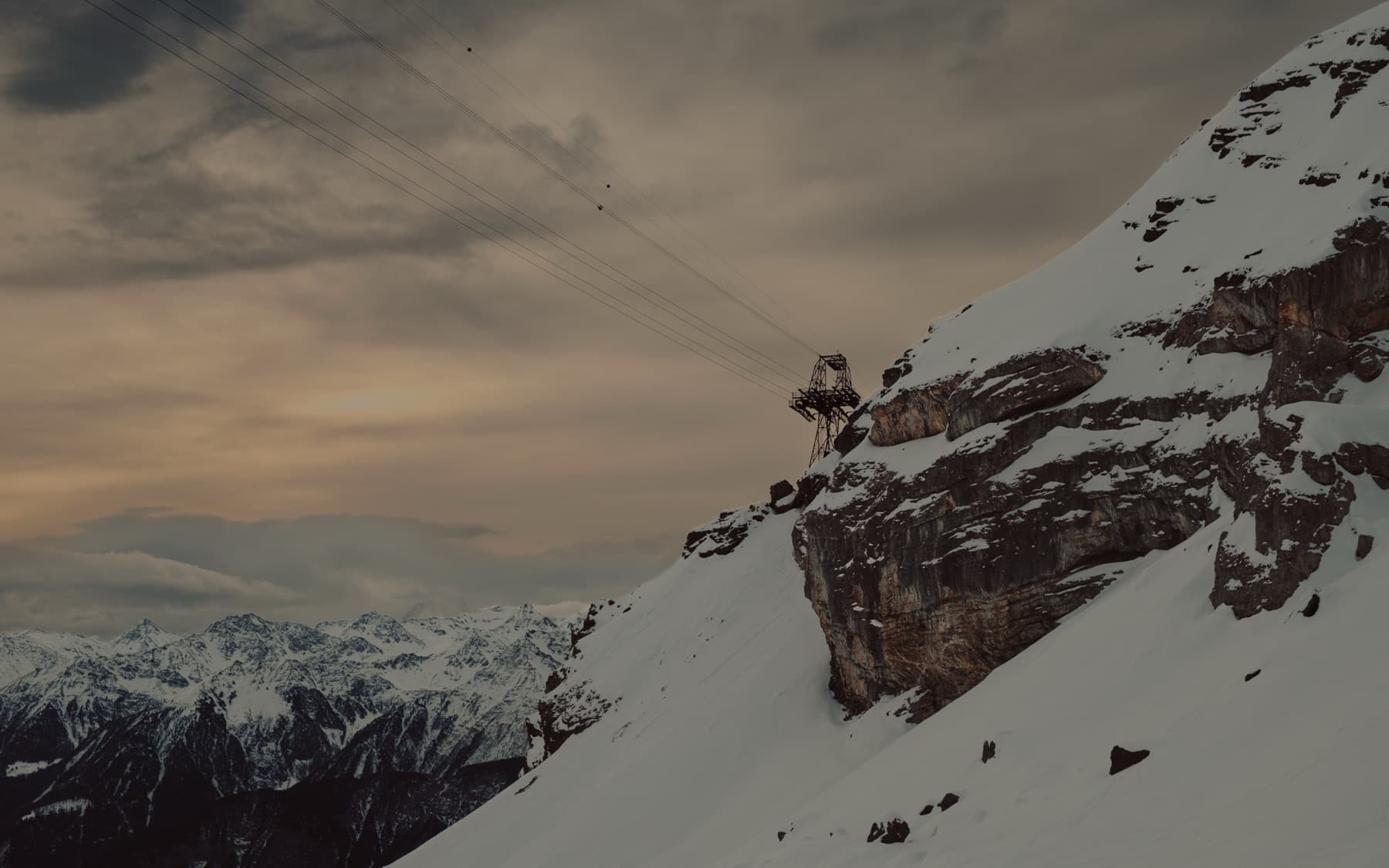 Cable car pylon perched on a steep snow-covered cliff edge with dramatic rock formations, distant alpine peaks and moody clouds filling the background
