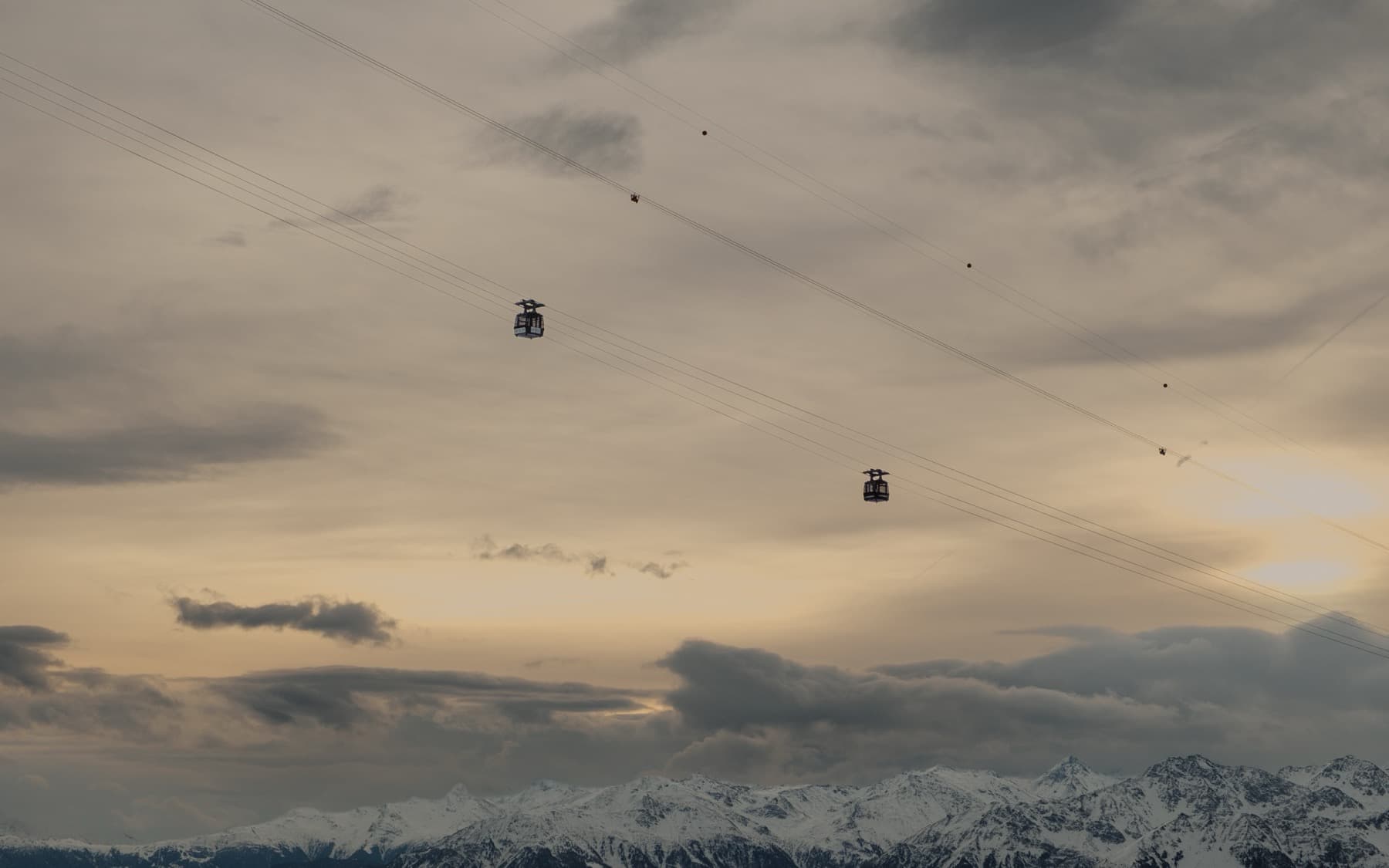 Two cable car gondolas suspended on wires against a dramatic cloudy sky at dusk, with a chain of snow-covered peaks along the horizon below