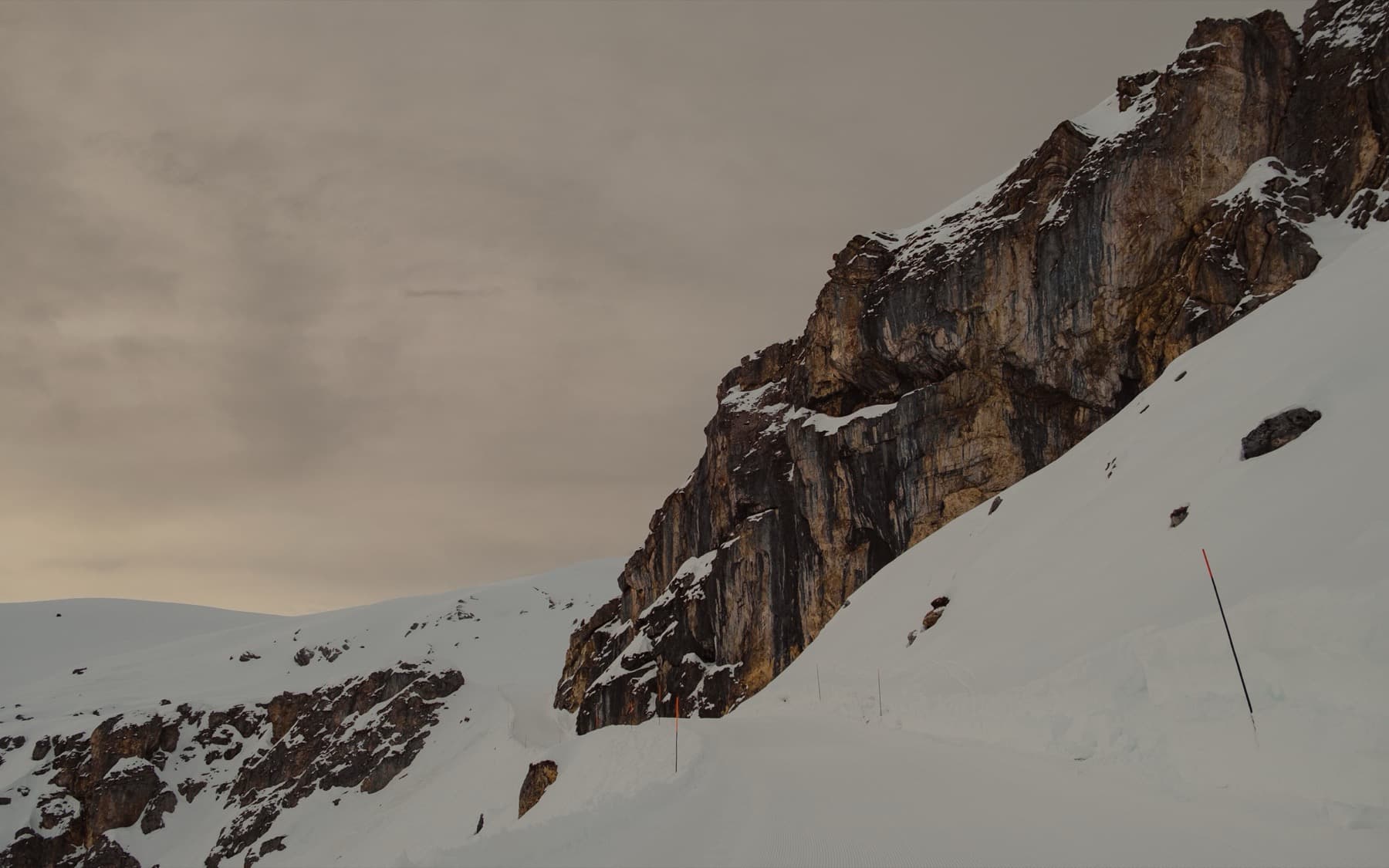 Massive exposed rock face rising from a snow-covered slope with orange piste markers, under a muted golden overcast sky