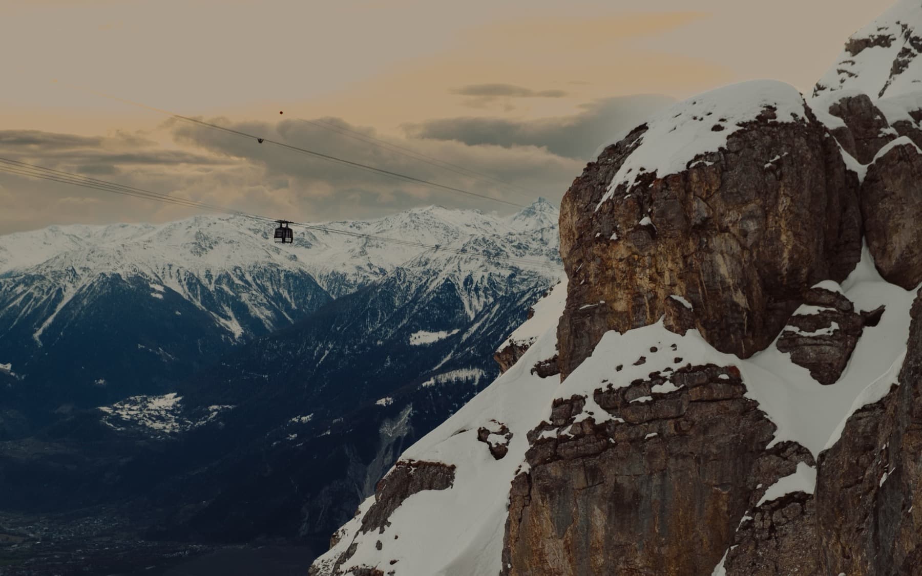 Cable car gondola silhouetted against a warm sunset sky, passing a dramatic snow-covered cliff face with exposed rock layers and distant peaks