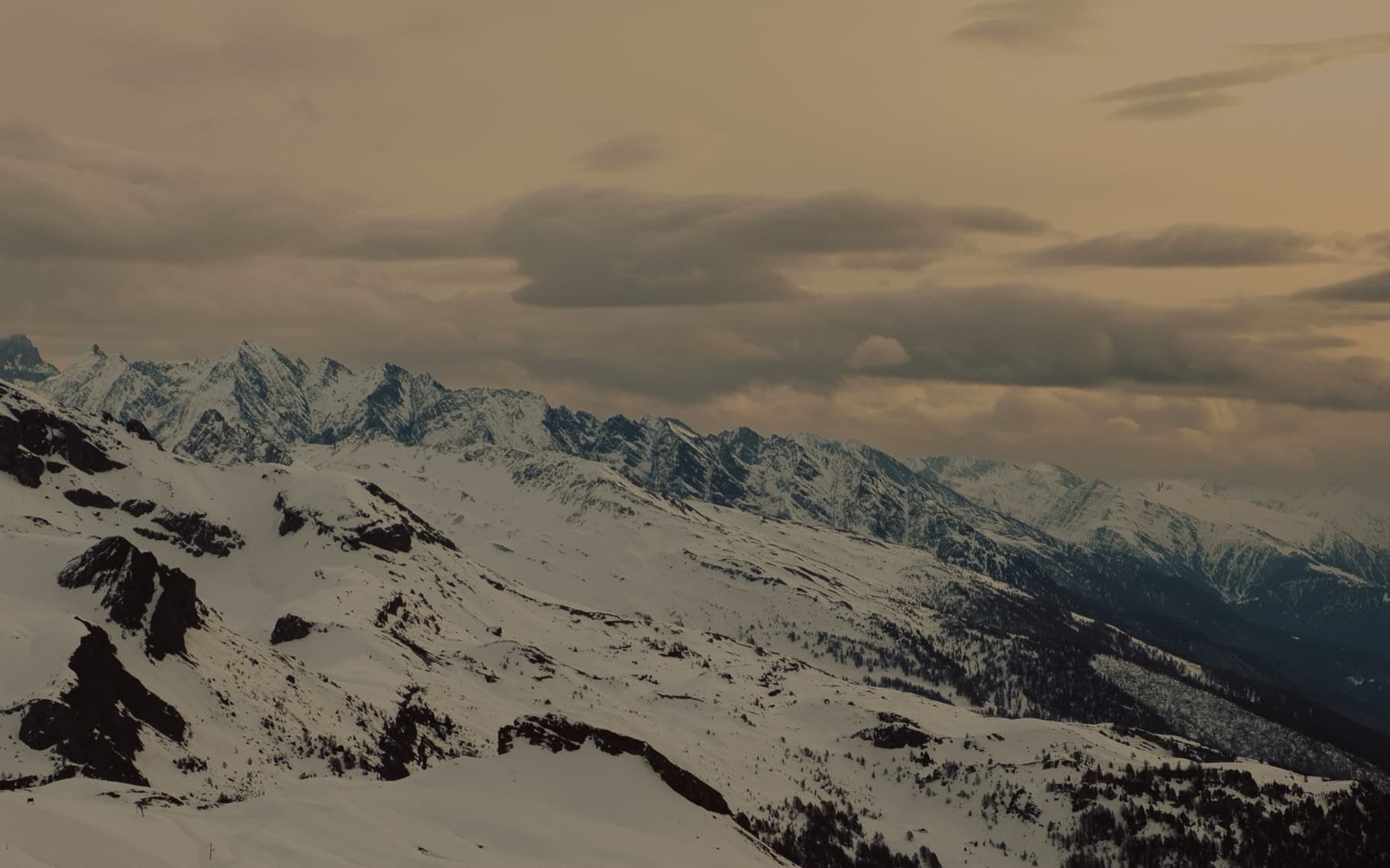 Wide panoramic view of snow-covered alpine ridges with rocky outcrops in the foreground, layered mountain peaks receding into a hazy overcast sky