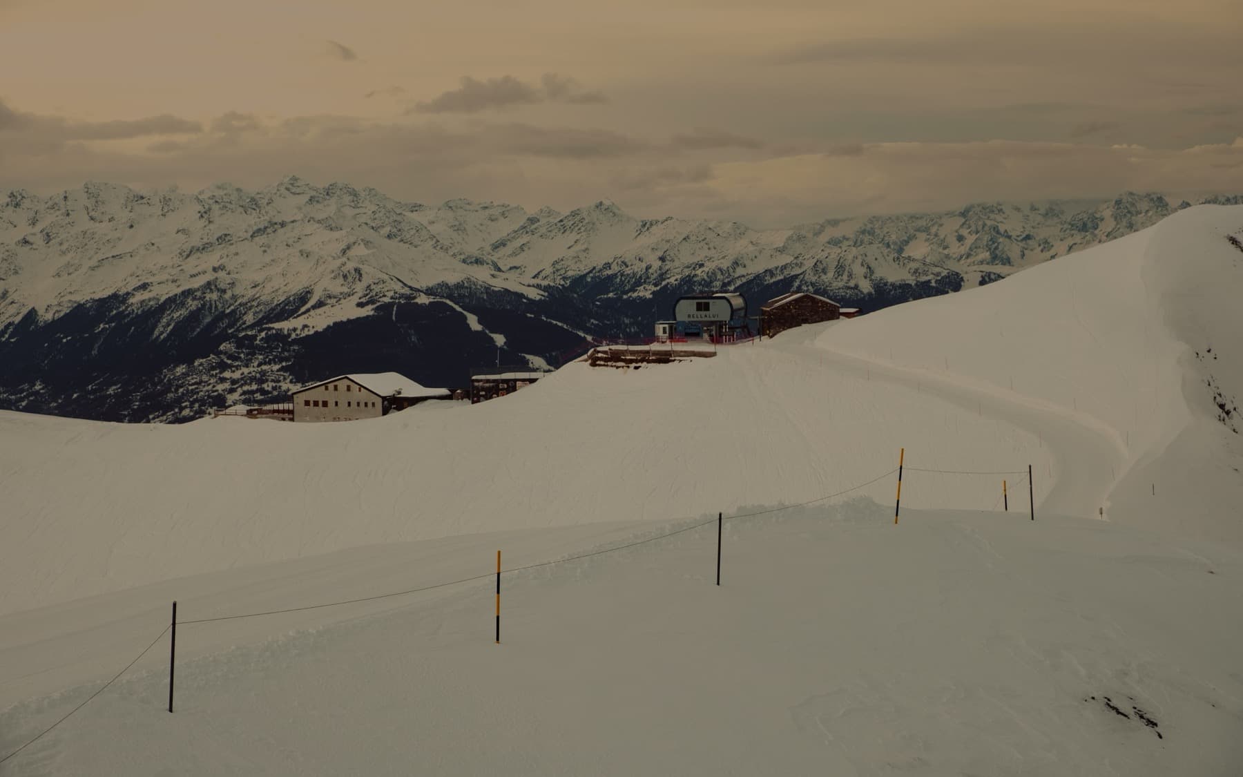 Mountain ski station buildings and chairlift on a snowy ridge with piste markers in the foreground, panoramic view of distant peaks under a golden overcast sky