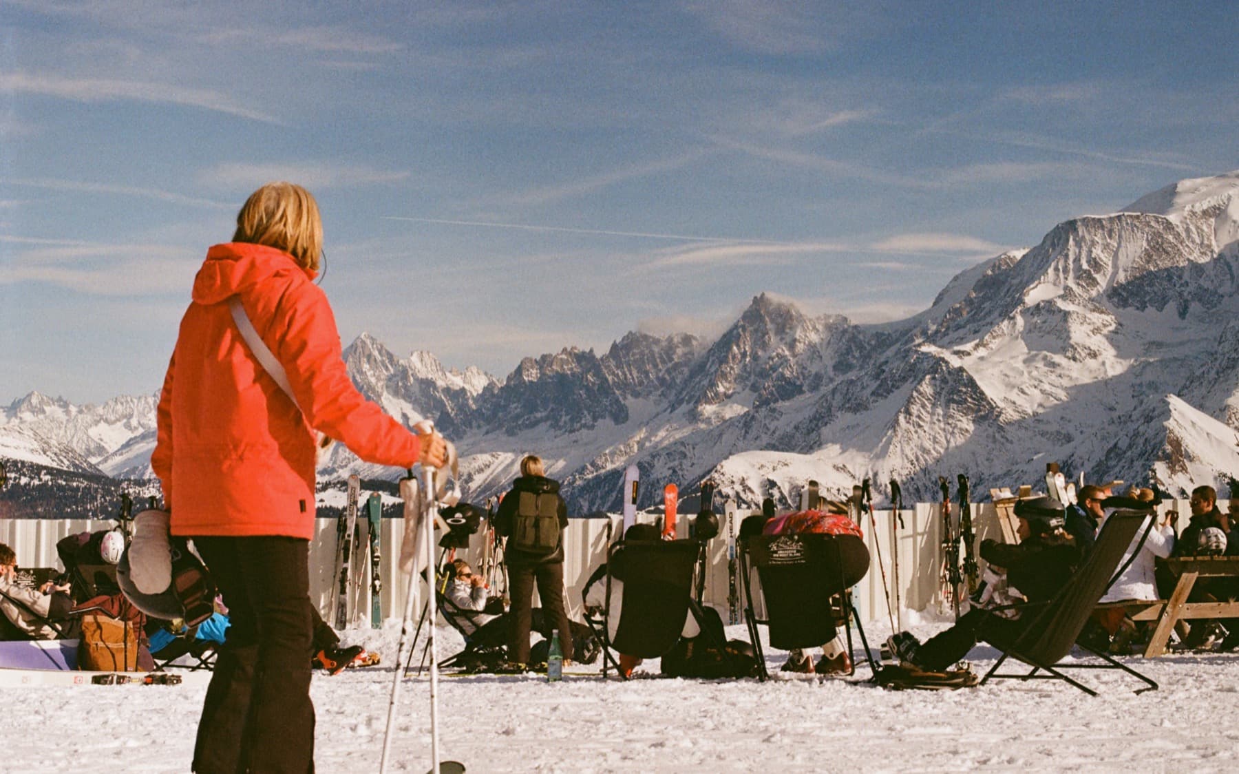 Skier in a red jacket standing with poles on a sunny mountaintop, facing a panoramic view of the Mont Blanc massif with other skiers lounging in deck chairs