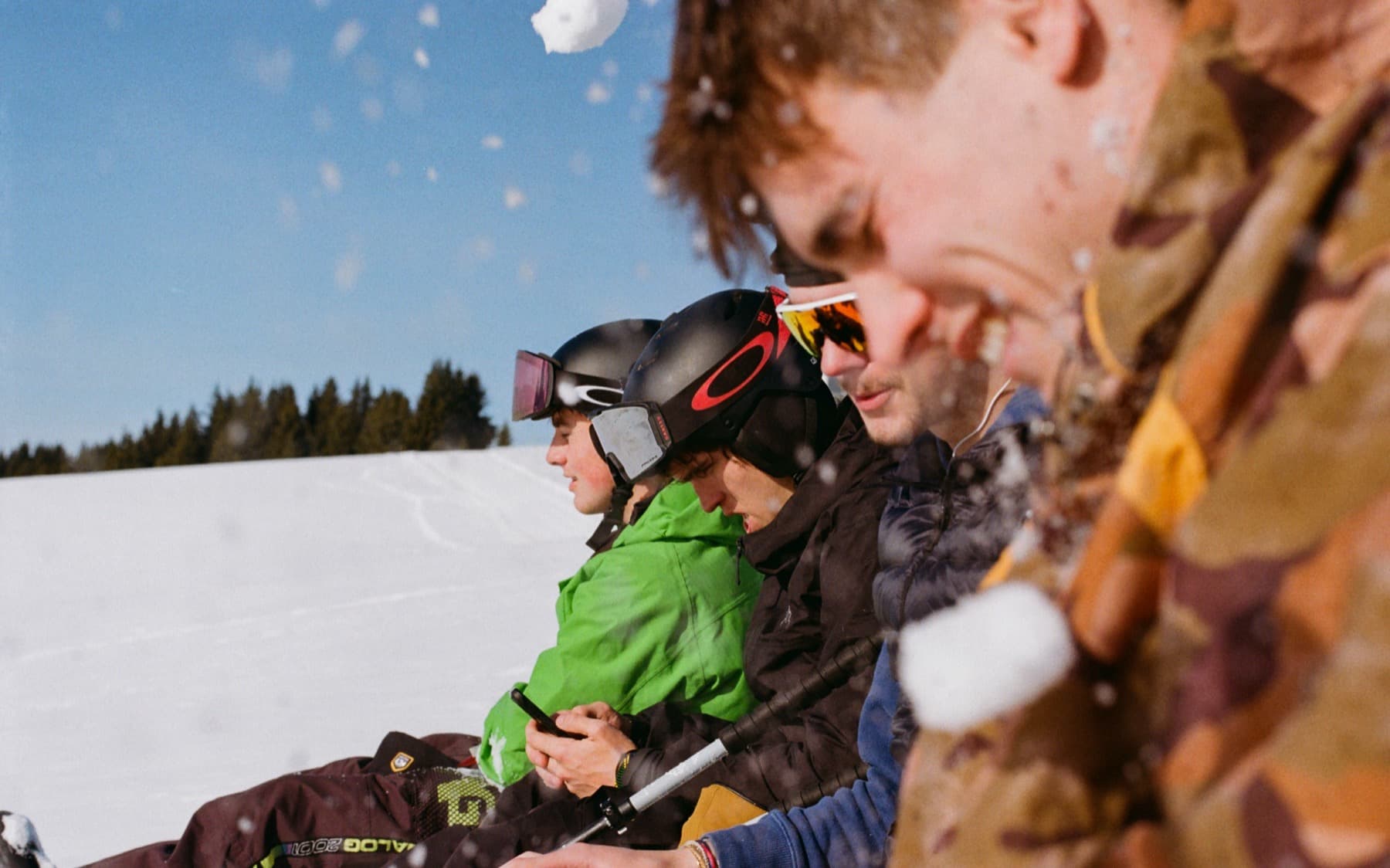 Group of friends in ski gear being hit by a snowball mid-air, laughing on a sunny slope with pine trees in the background