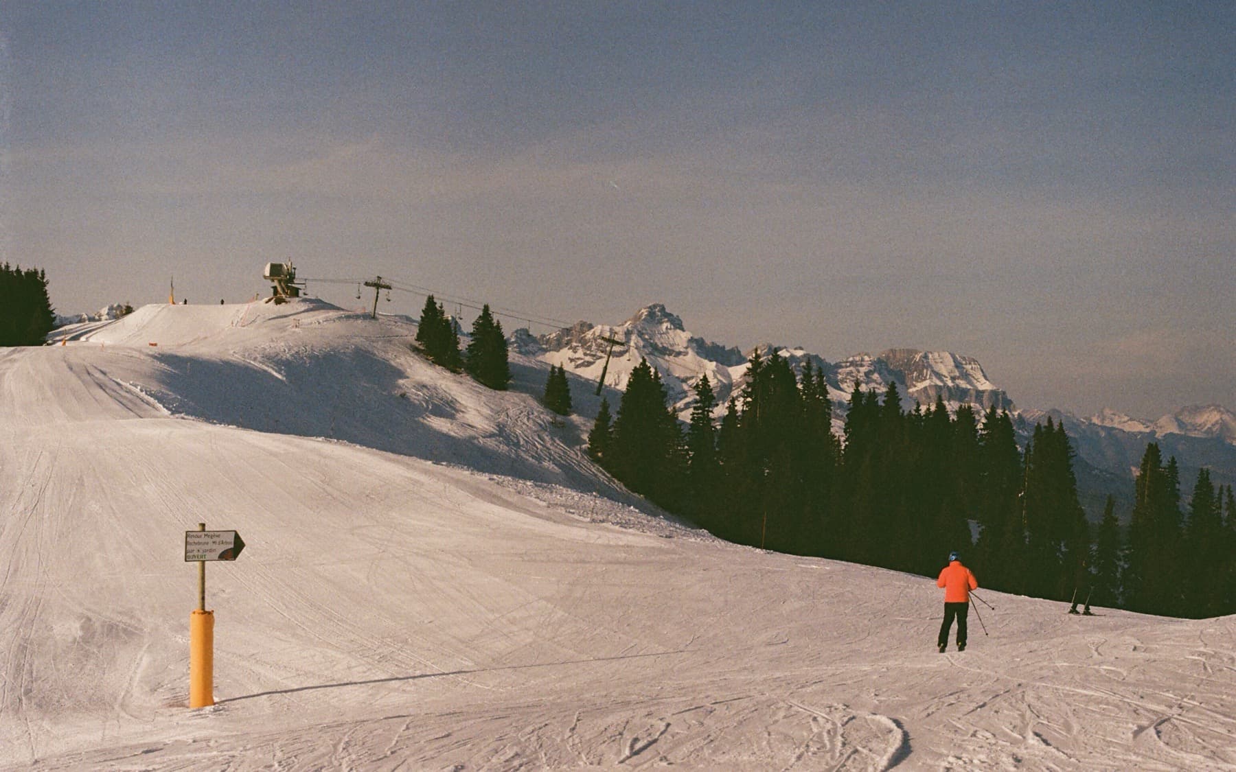 Lone skier in an orange jacket gliding down a groomed piste toward a chairlift station, with pine trees and distant peaks in late afternoon light
