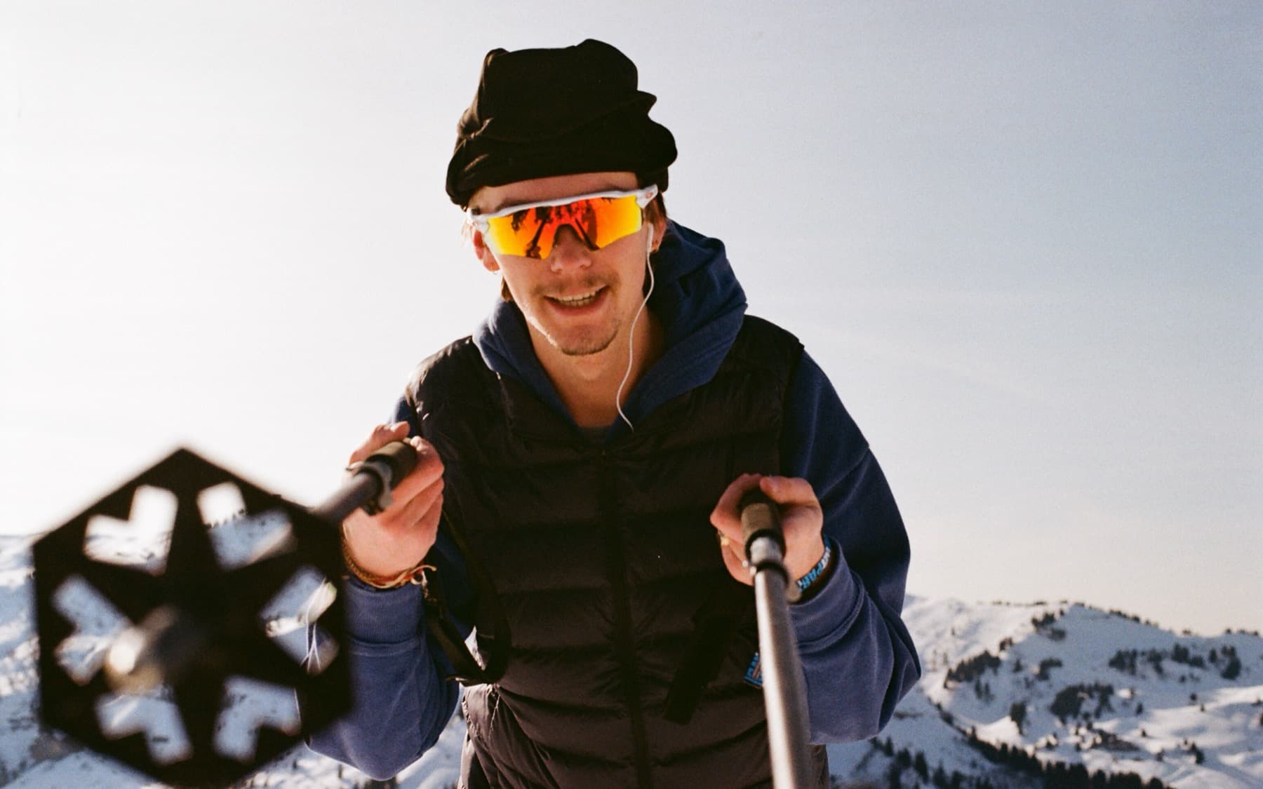 Close-up portrait of a smiling skier wearing orange mirrored sunglasses and a dark puffer jacket, holding ski poles with snowy mountains behind