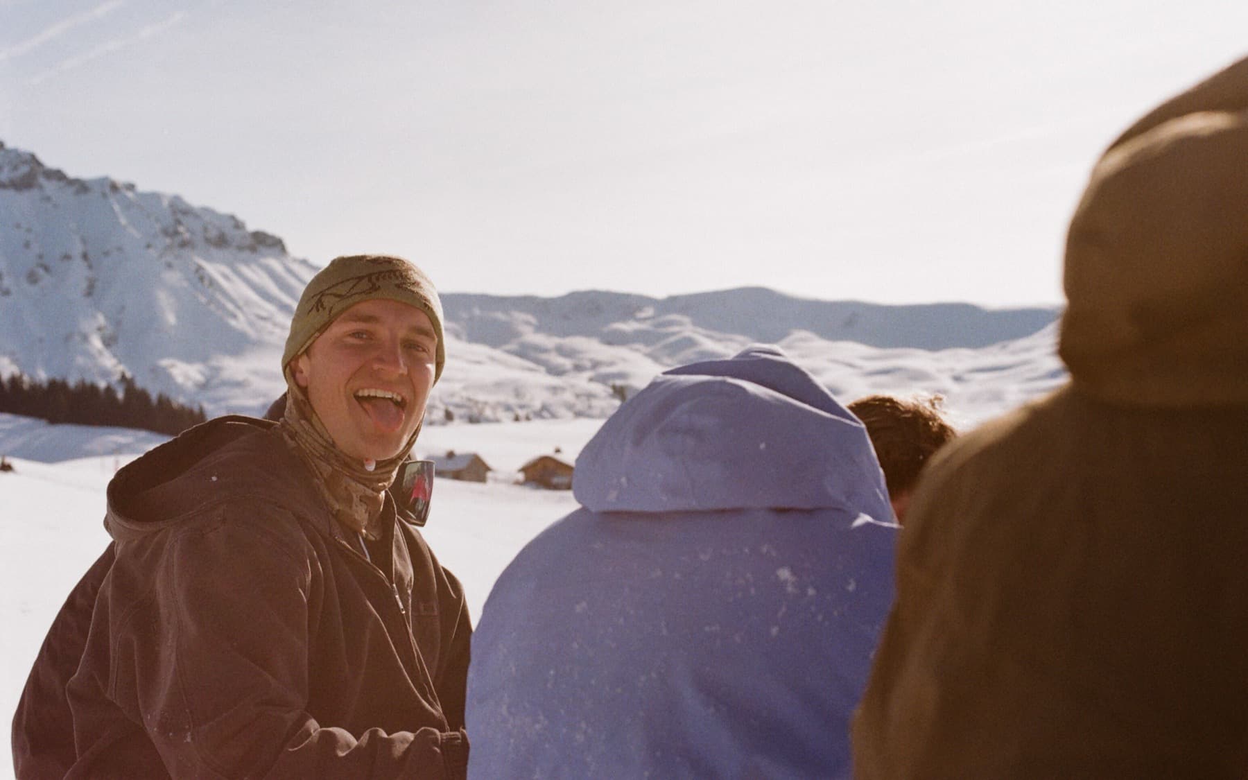Young man in a beanie laughing among friends on a snowy plateau, with alpine chalets and snow-covered hills in the warm afternoon sun