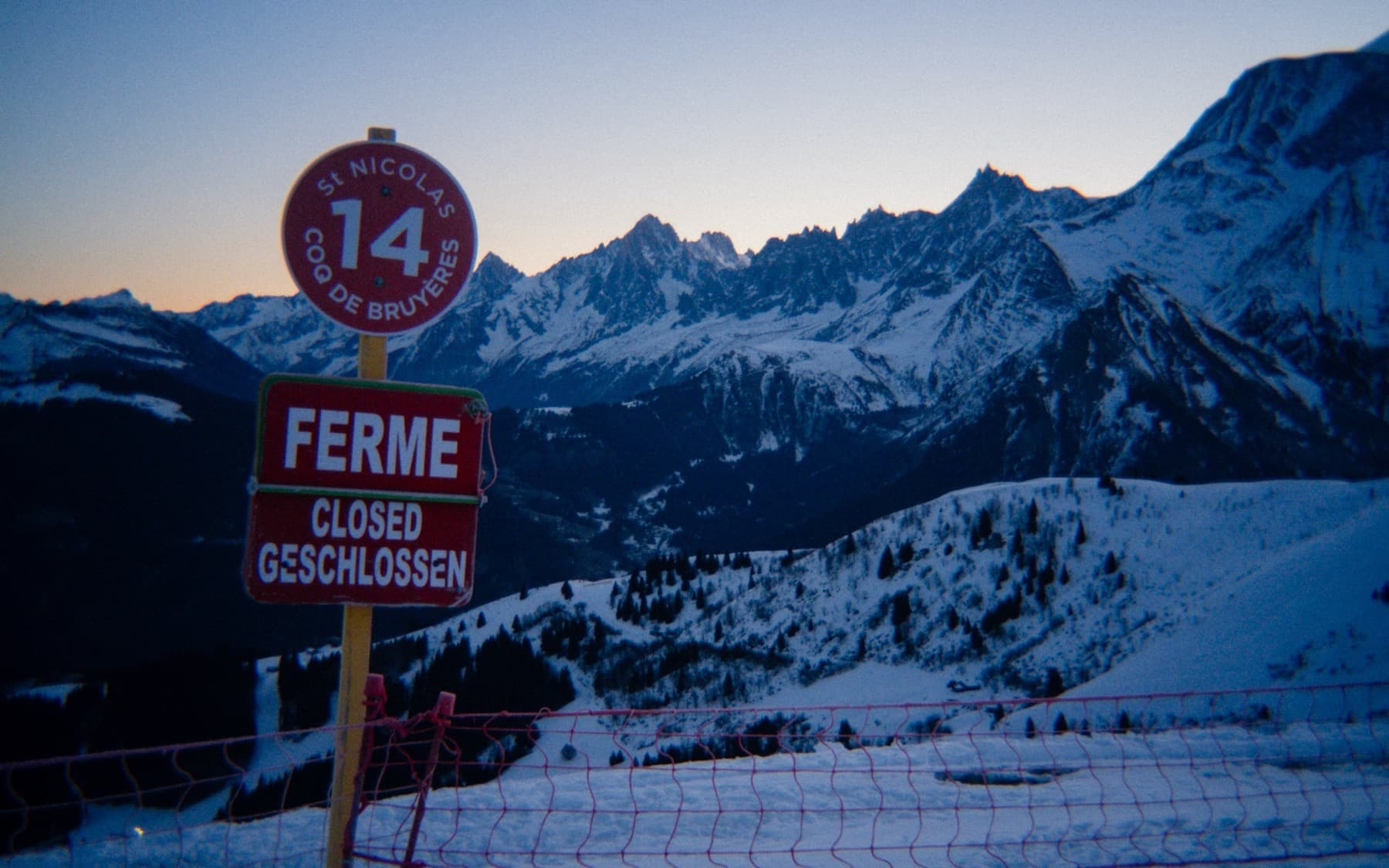 Closed ski run sign reading Ferme at dusk with dramatic snow-capped peaks and deep valleys in the fading twilight behind