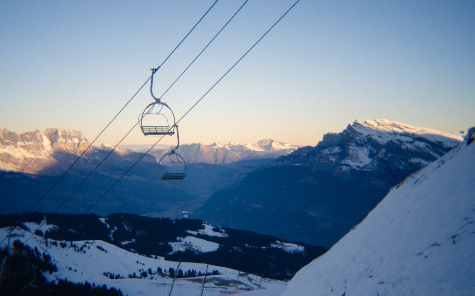 Empty chairlift silhouetted against a golden dusk sky, suspended above snow-covered slopes with alpine peaks glowing in the distance