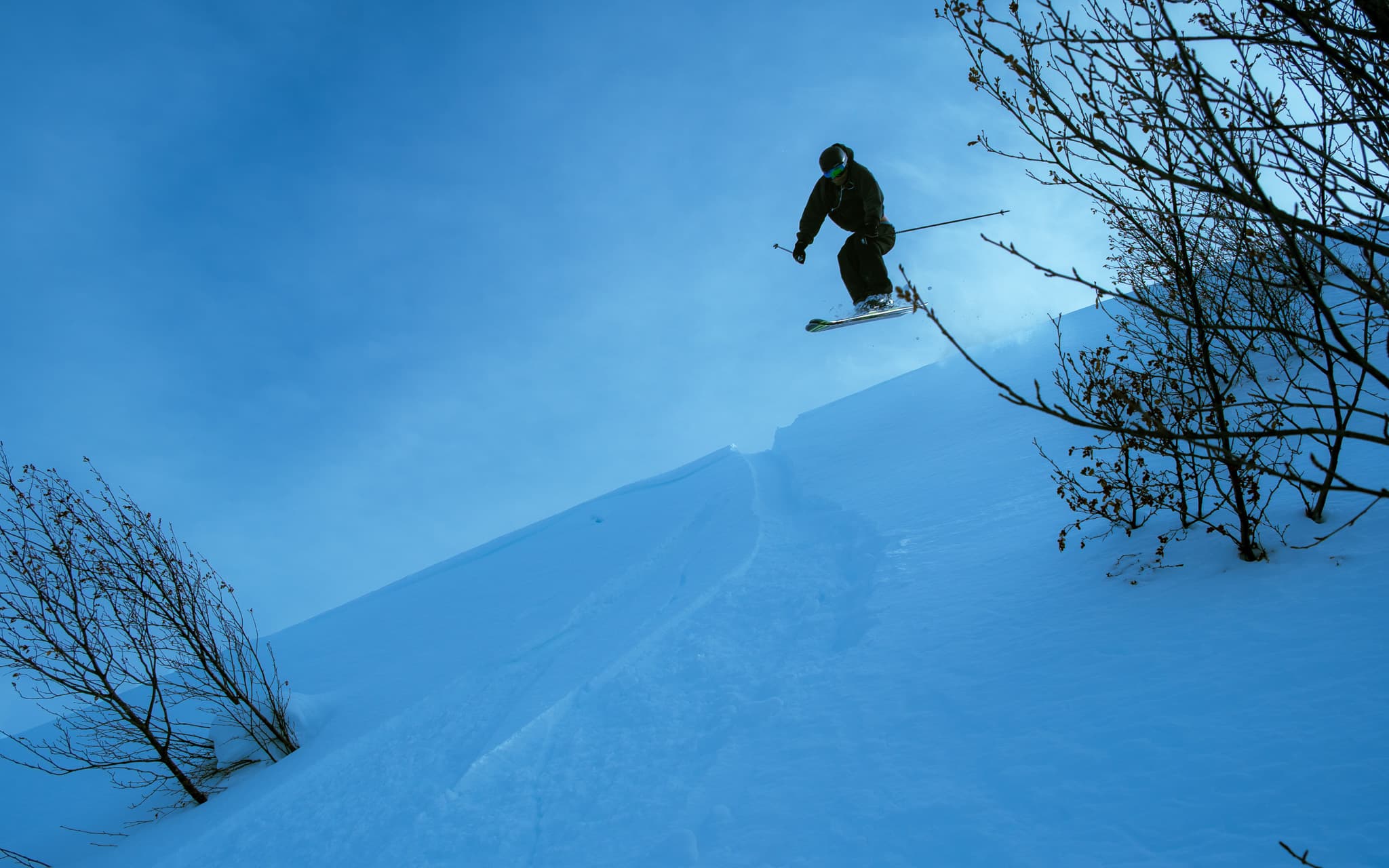 Skier mid-jump above a powder snow drop with ski poles extended against a clear blue sky