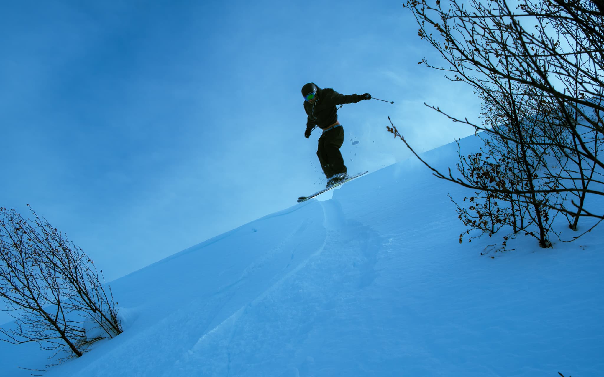 Skier at the peak of a jump off a snow lip with bare branches and blue sky behind