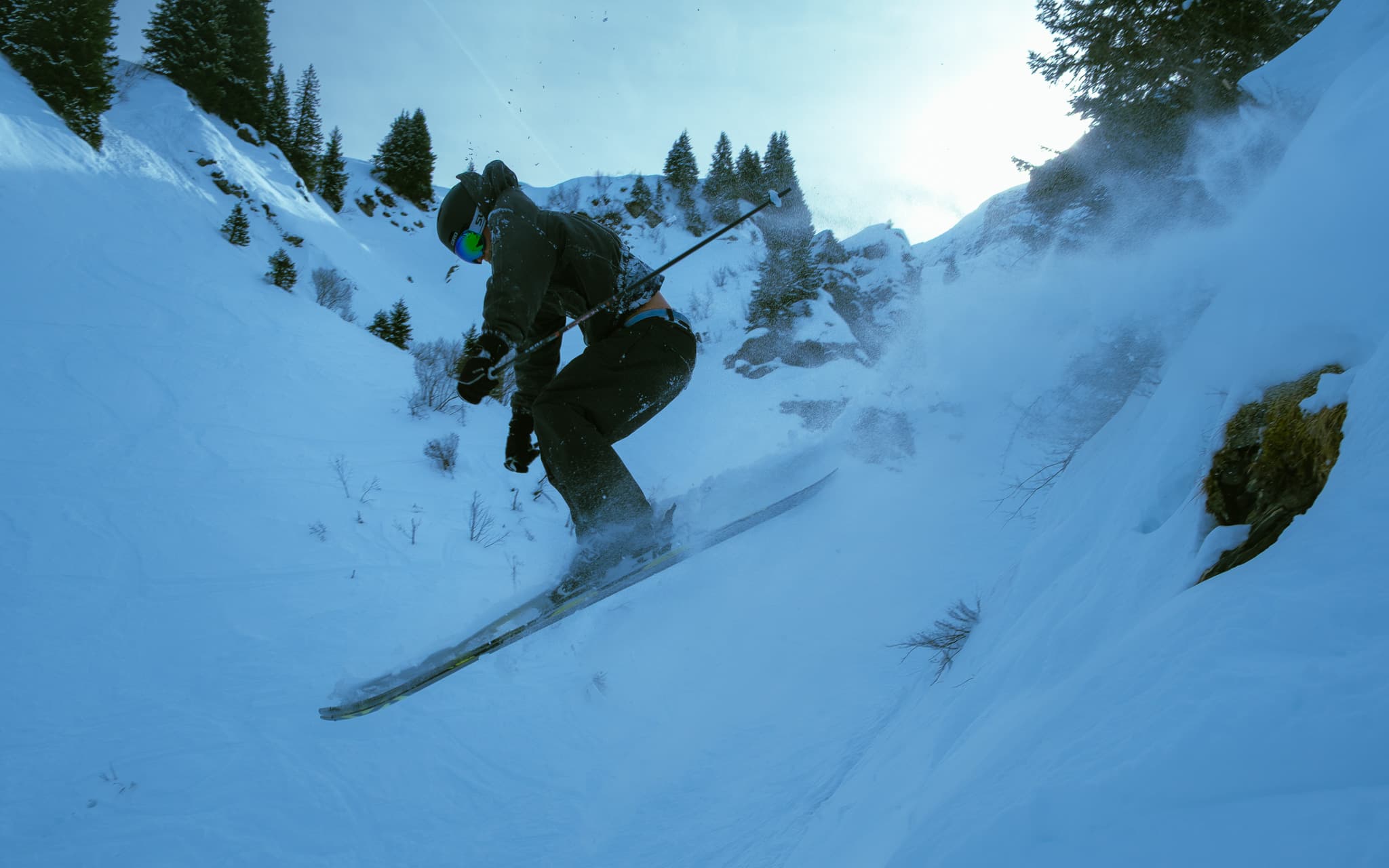 Skier landing a drop through powder snow between rocks with pine trees and mountain peaks in the background