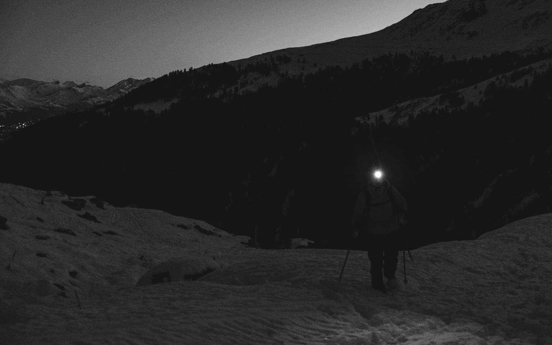 Hiker with a headlamp walking through deep snow at night in a mountain valley, dark forested slopes rising on either side
