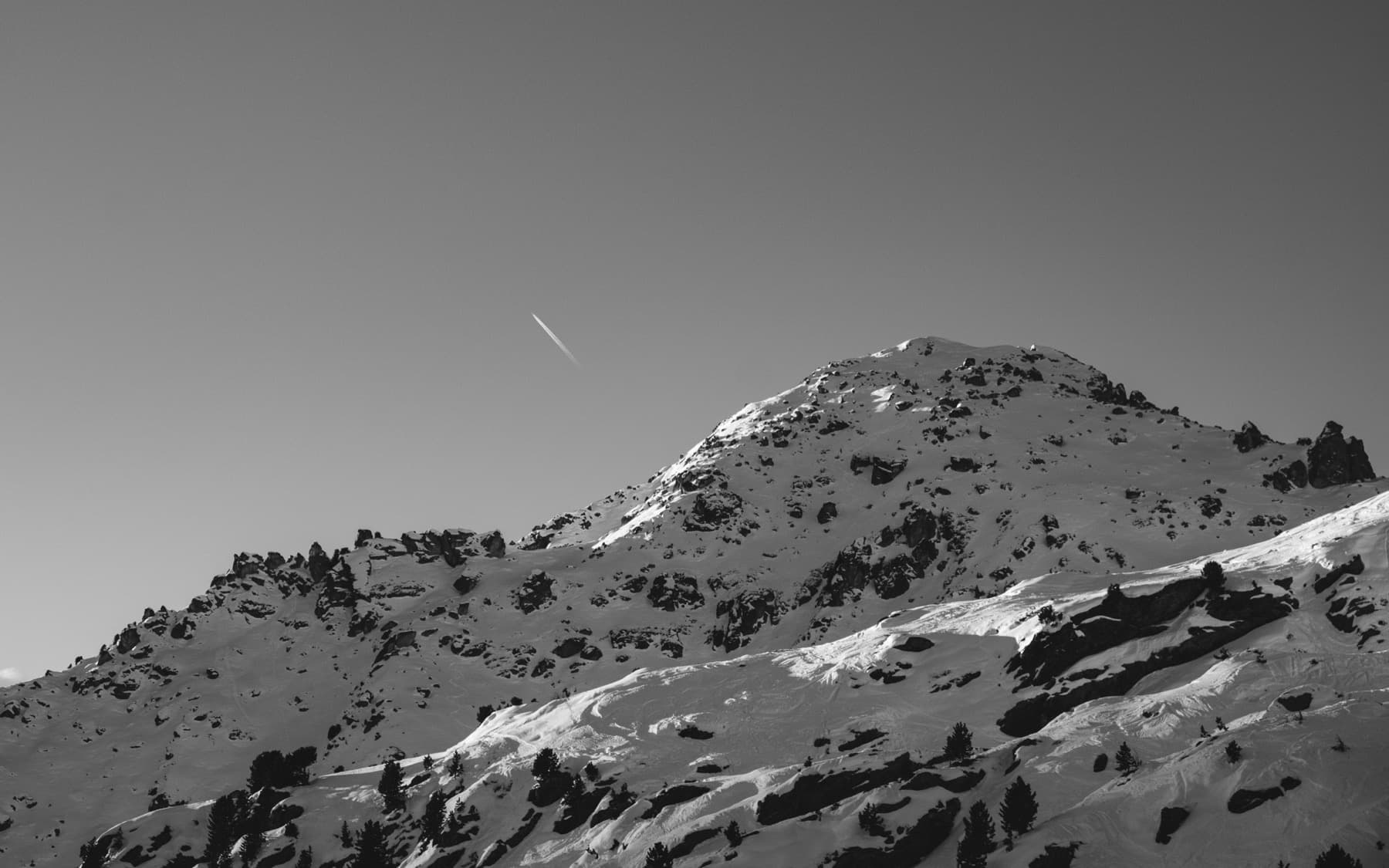 Snow-dusted mountain peak with a jet contrail streaking across a clear sky above, scattered trees on the lower slopes, shot in black and white