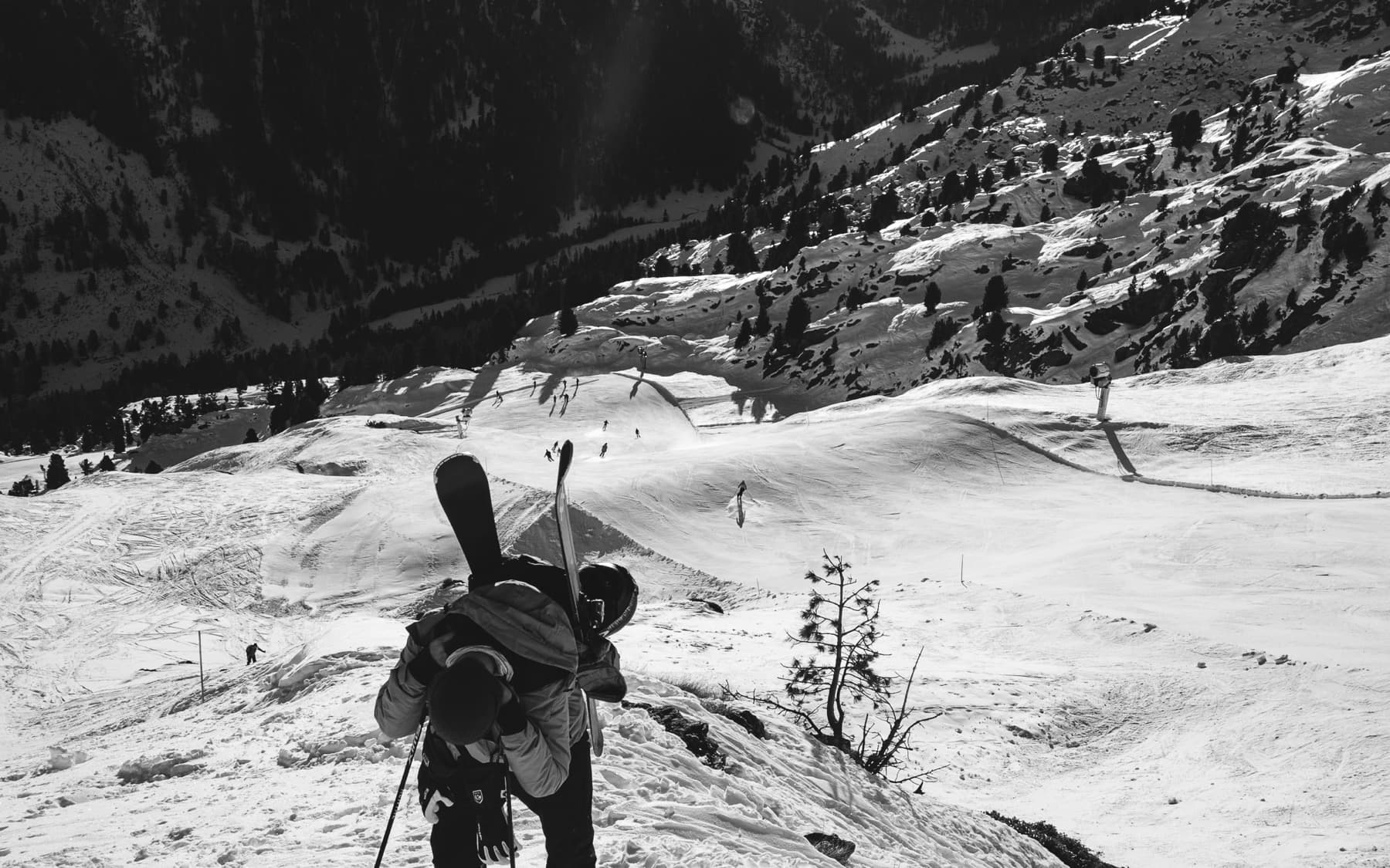 Ski tourer carrying skis on a backpack, hiking down a snowy ridge overlooking a valley with other skiers below, shot in black and white