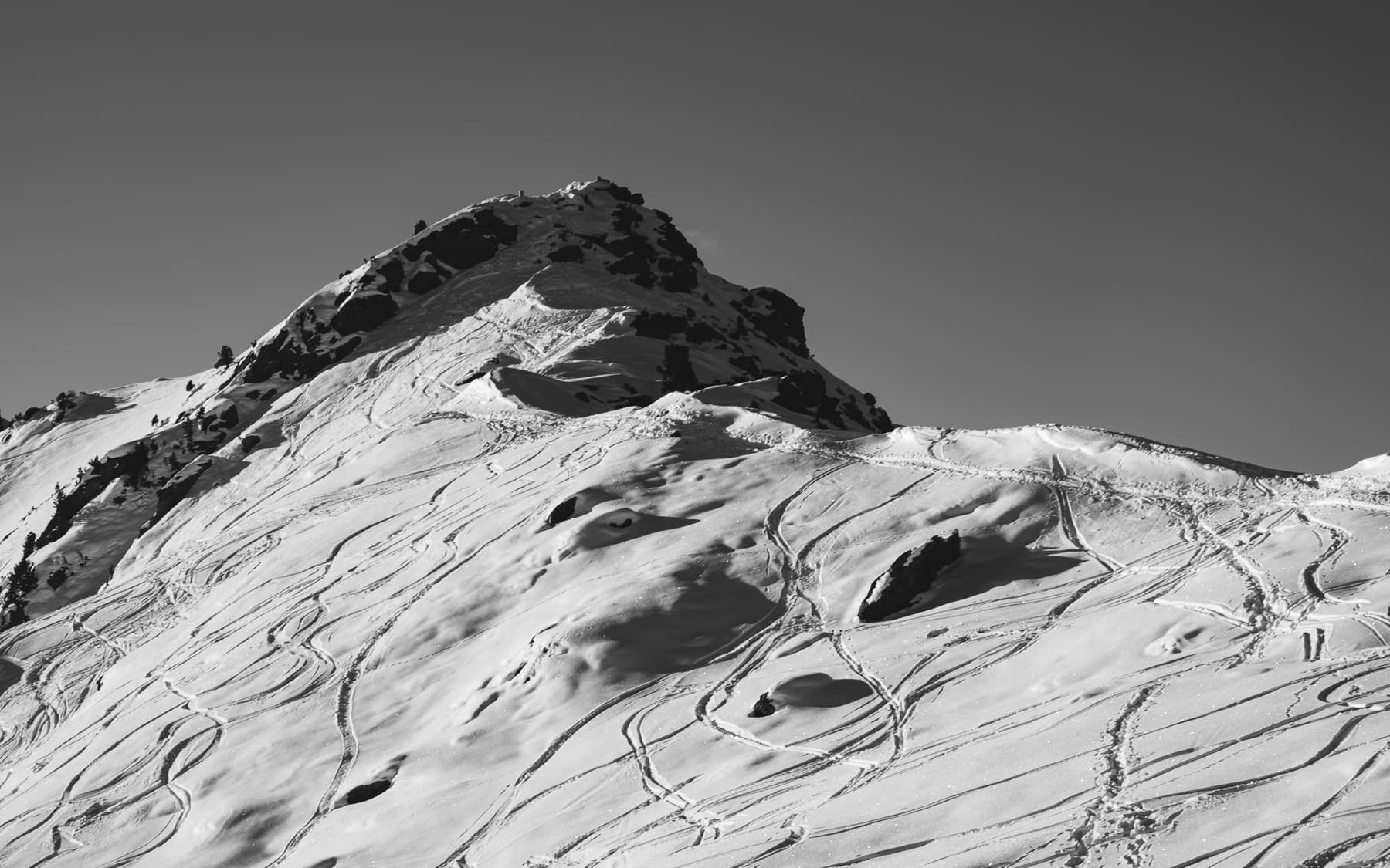 Mountain peak covered in ski tracks weaving through fresh snow, with exposed rocky summit, shot in high-contrast black and white