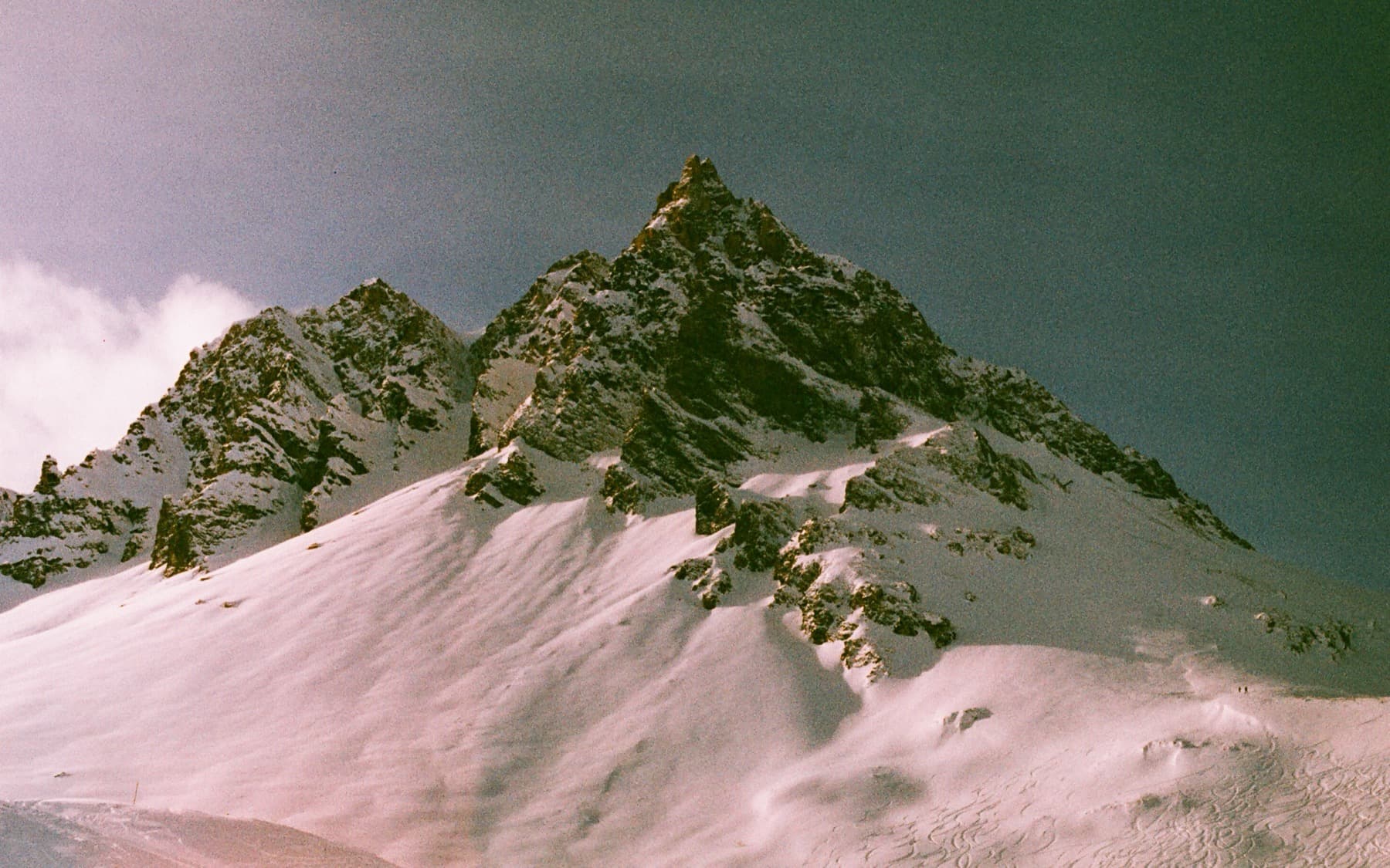 Sharp rocky summit with steep snow-filled couloirs and ski tracks visible on the lower slopes under a deep teal sky