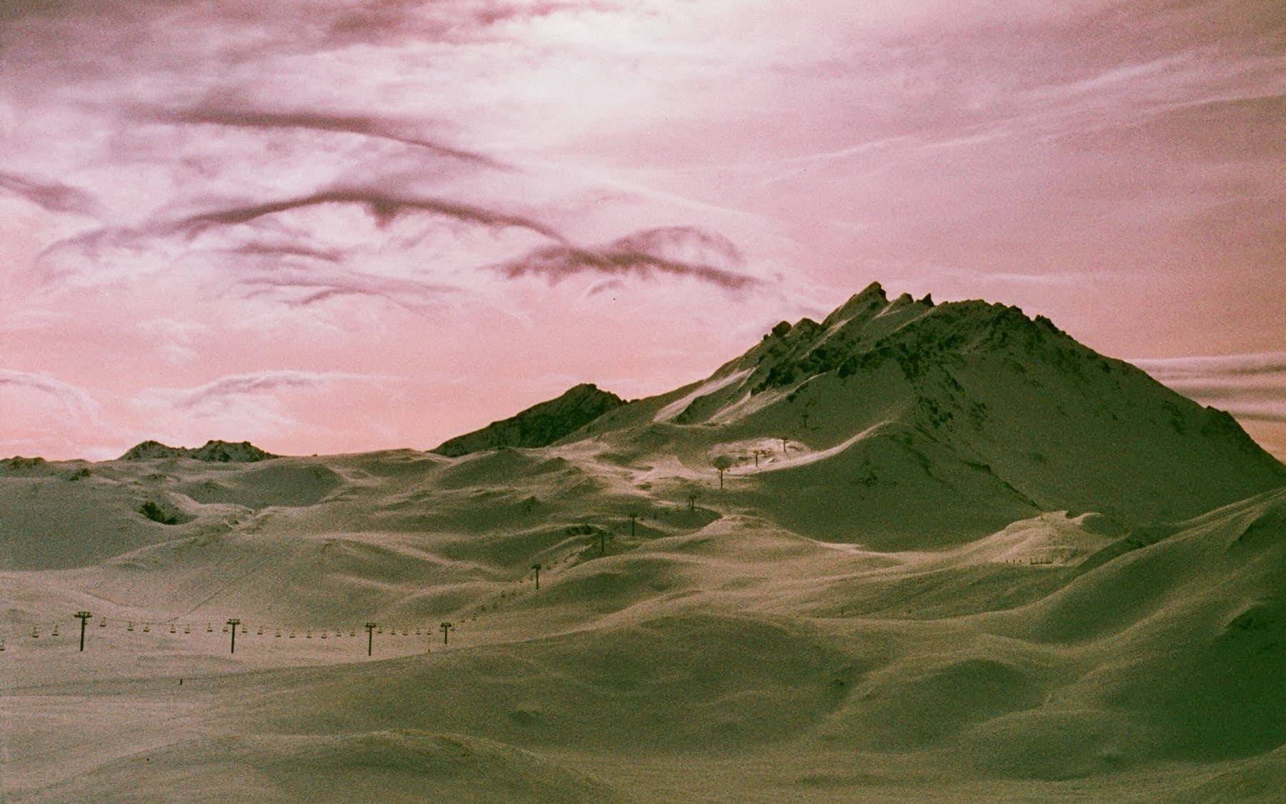 Rocky mountain peak rising above undulating snow-covered terrain with ski lift poles visible, shot on expired film giving a warm pink-toned sky