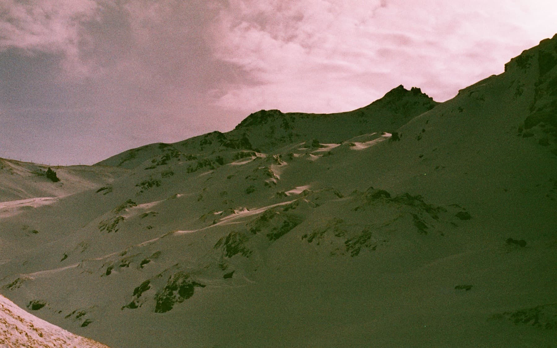 Rugged mountainside with patches of snow and exposed rock under a hazy pink sky, shot with warm expired film tones